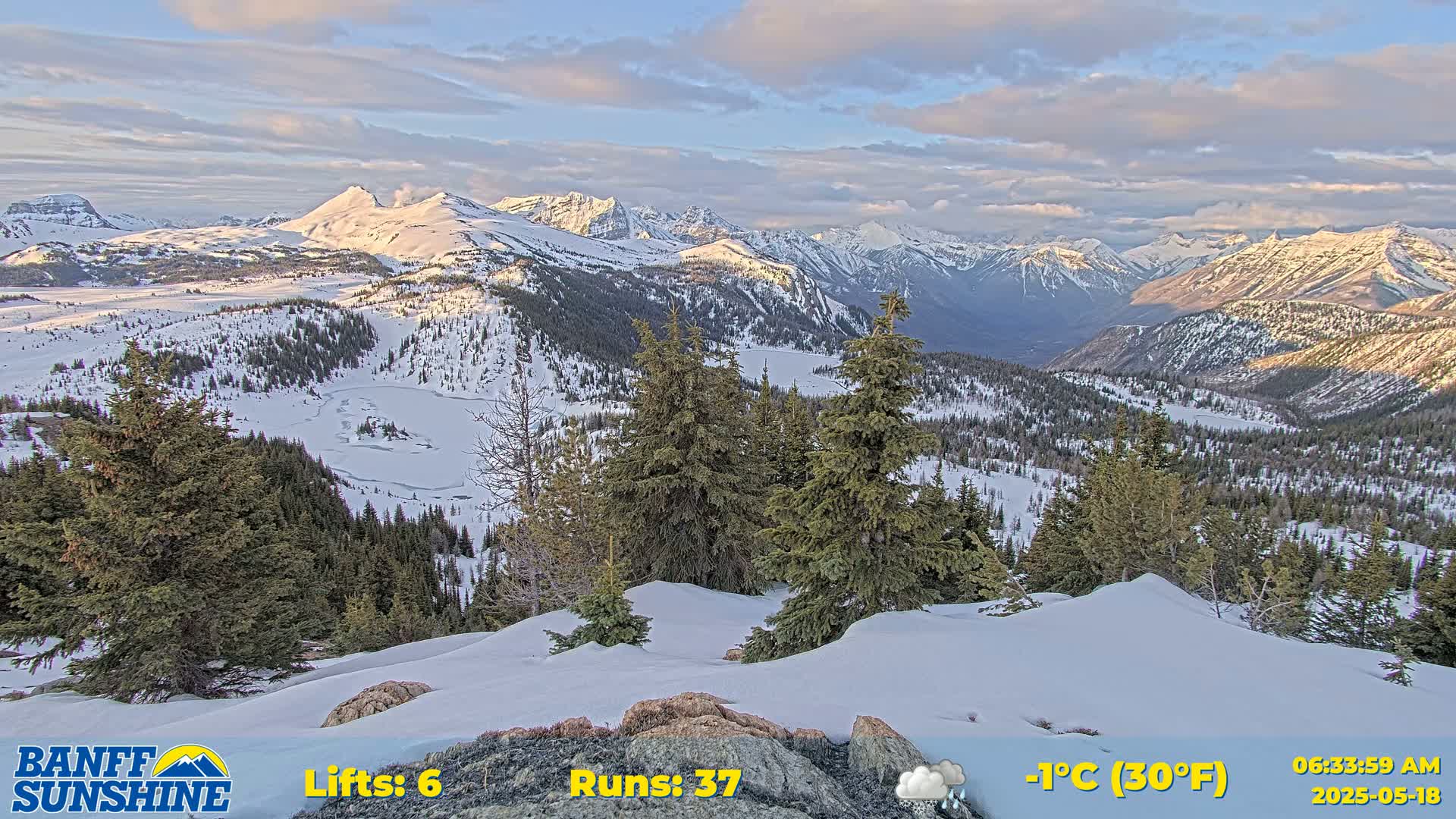 A snow-covered mountain landscape with evergreens under a partly cloudy sky shows a valley below and a partly frozen lake.