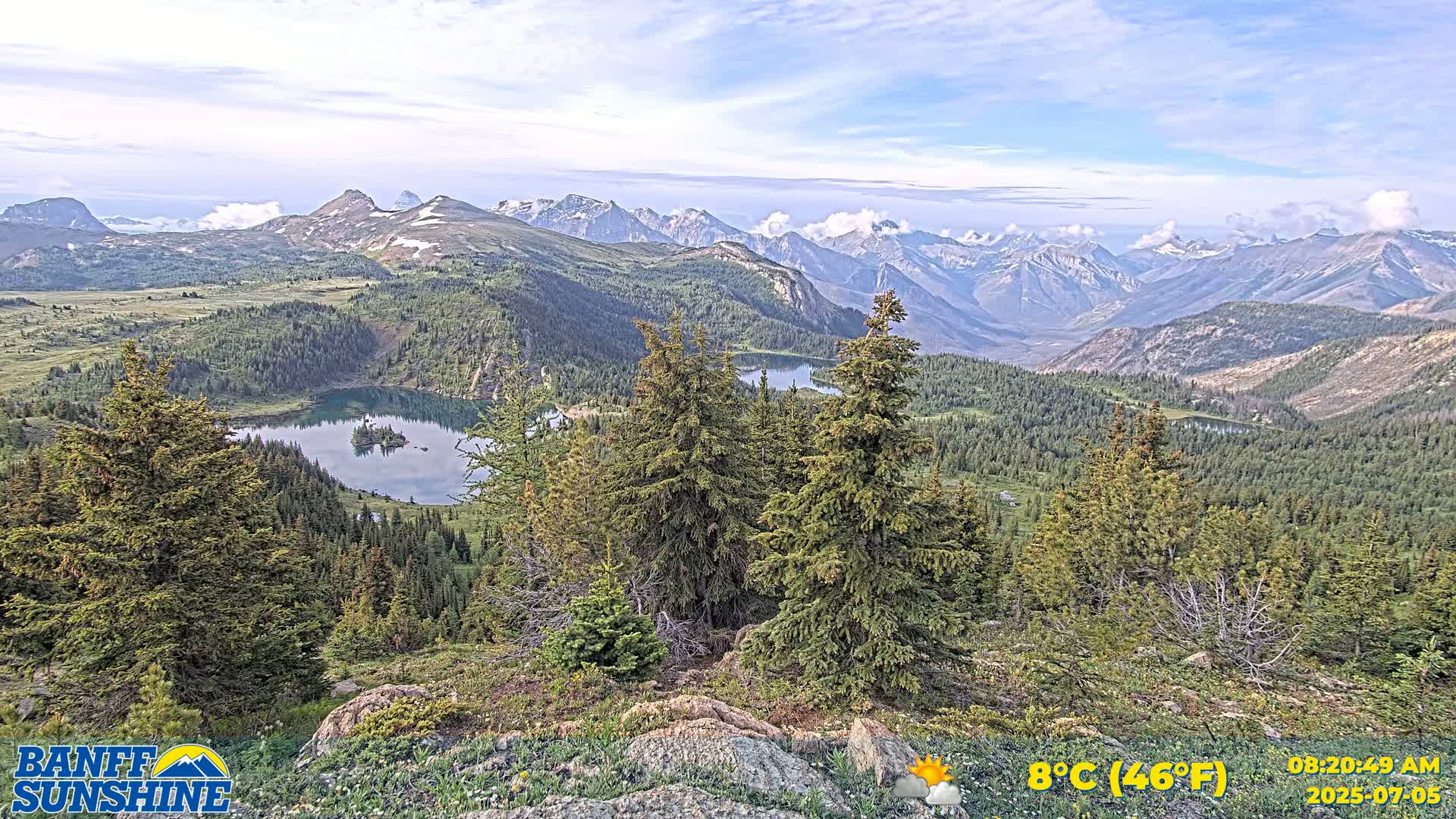 A mountain vista shows a valley of evergreen trees and a lake with a small island, under partly cloudy skies.