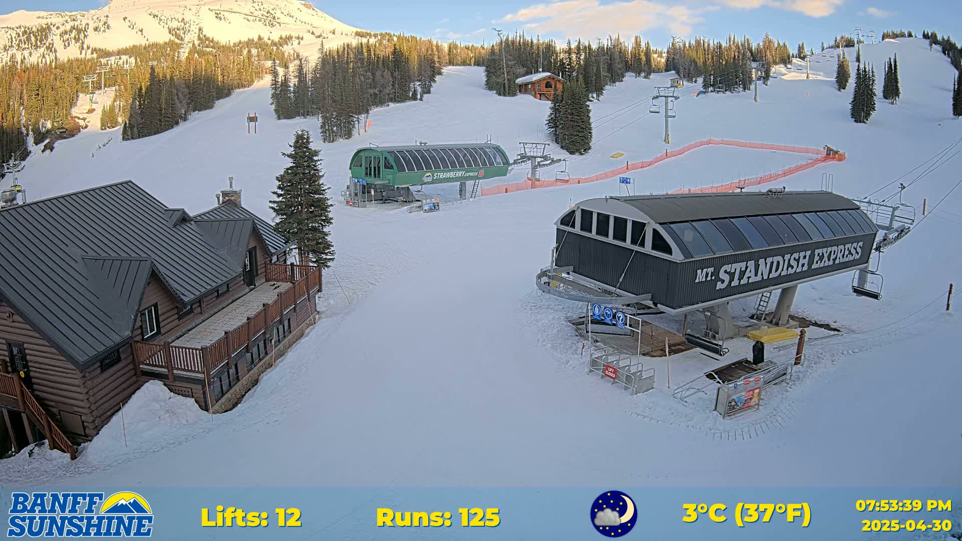 A snow-covered ski resort with two chairlifts, a log cabin, and a mountain in the background under a partly cloudy sky.