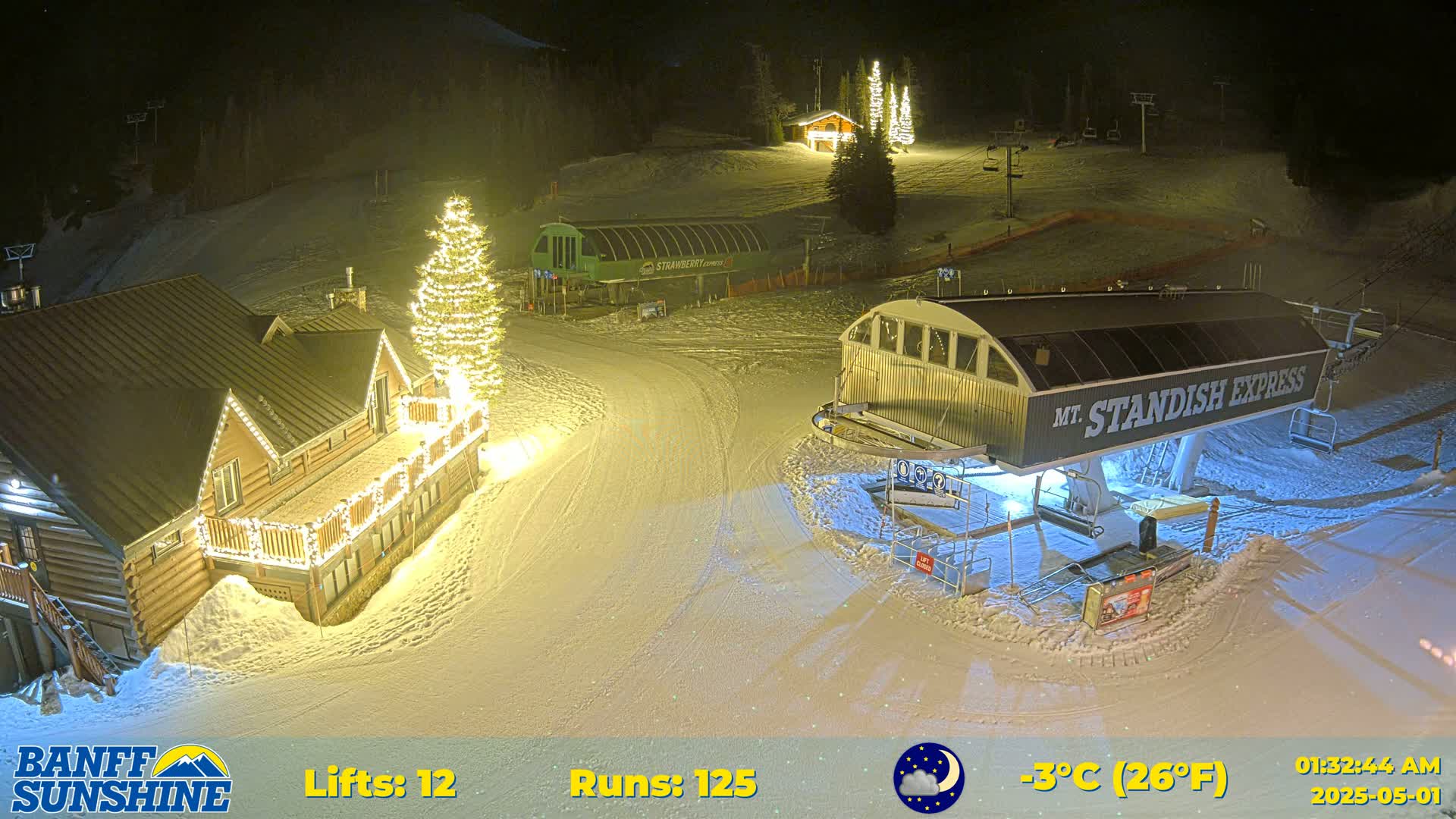 A nighttime view of a snow-covered ski resort with illuminated buildings and a ski lift, under a clear night sky with a temperature of -3°C (26°F).