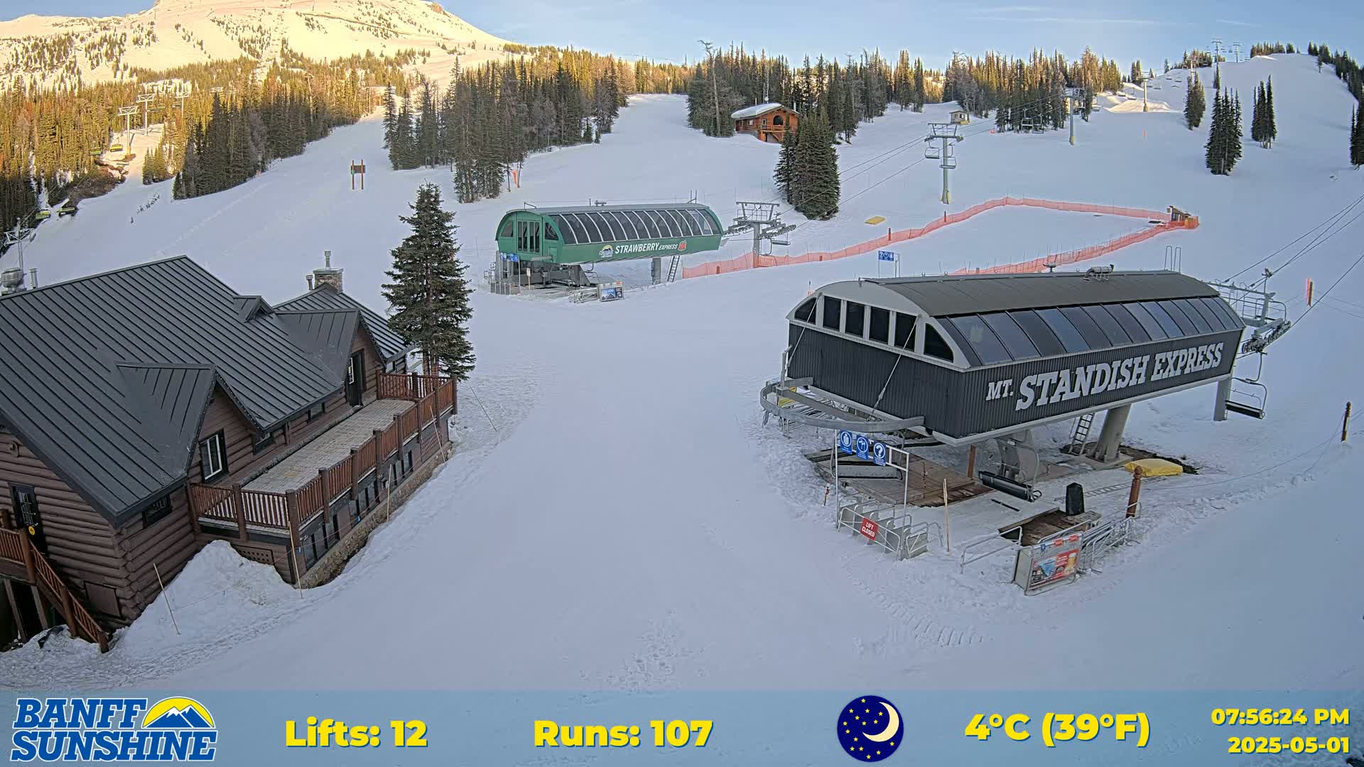 A snow-covered ski resort with two chairlifts, a log cabin, and a mountain in the background under a clear, twilight sky.