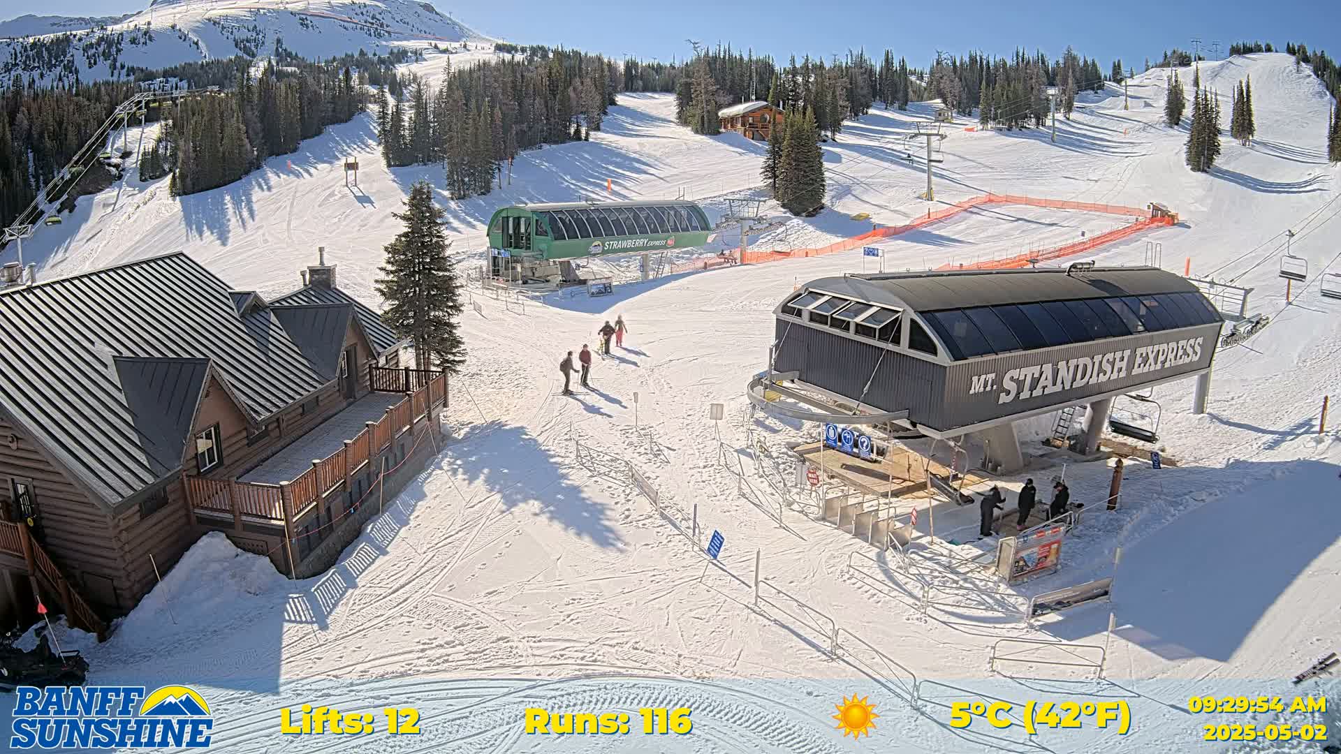 A sunny ski resort with two chairlifts, a lodge, and several skiers on a snow-covered mountain.