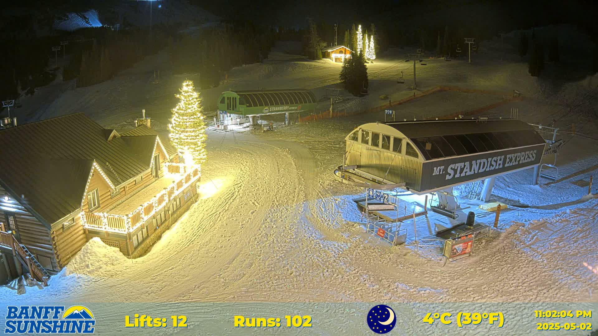 A nighttime view of a snow-covered ski resort with illuminated buildings and ski lifts, under a clear sky with a temperature of 4°C (39°F).