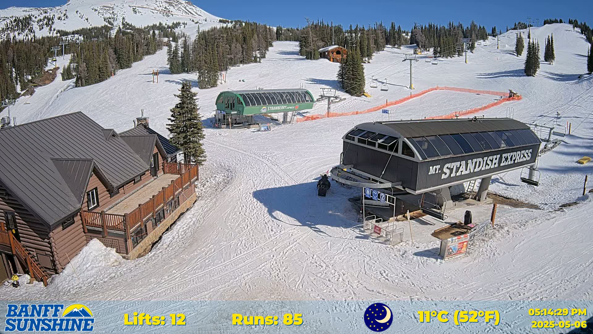 A snow-covered ski resort on a sunny day shows two chairlifts, a log cabin, and several ski runs.