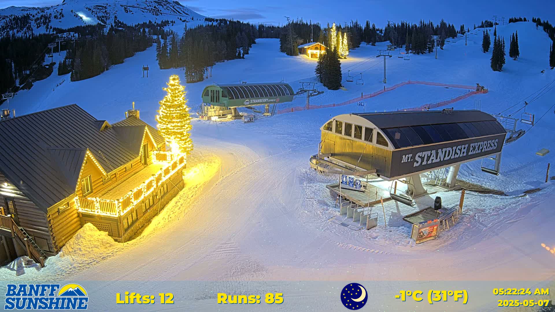 A snow-covered ski resort at night, illuminated by lights on buildings and a Christmas tree, under a twilight sky.