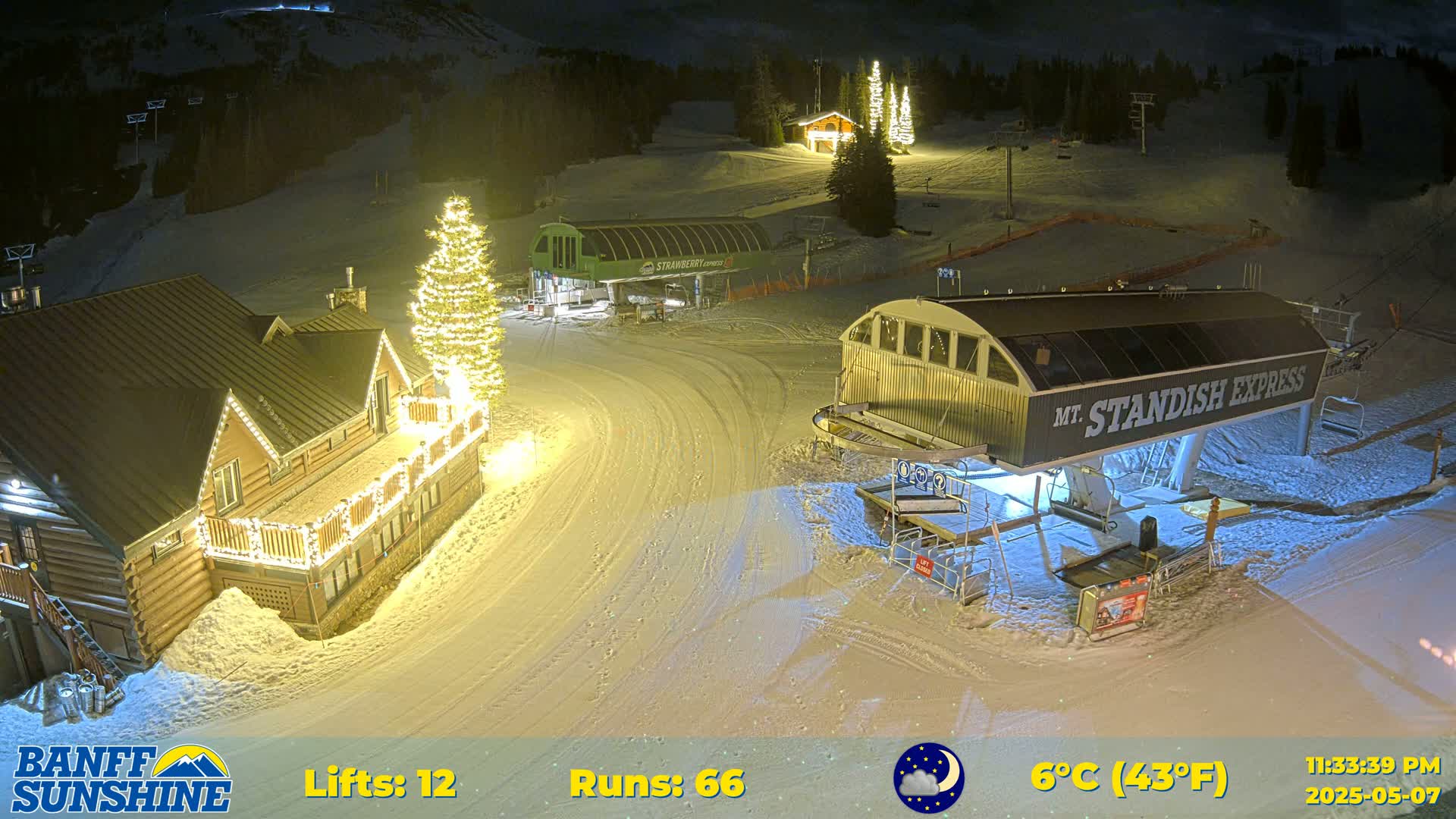 A nighttime view of a snow-covered ski resort with illuminated buildings and ski lifts, under a partly cloudy sky and a temperature of 6 degrees Celsius.
