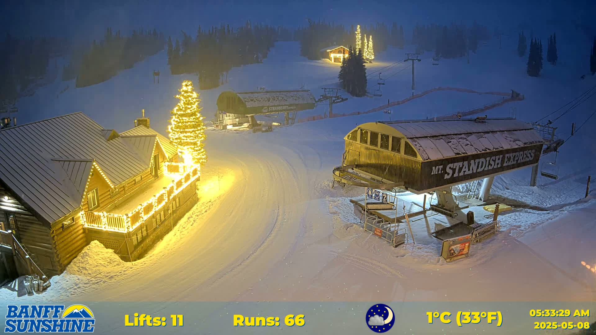 A snow-covered ski resort at night, illuminated by lights on buildings and a Christmas tree, with a partly cloudy sky and a temperature of 1°C (33°F).
