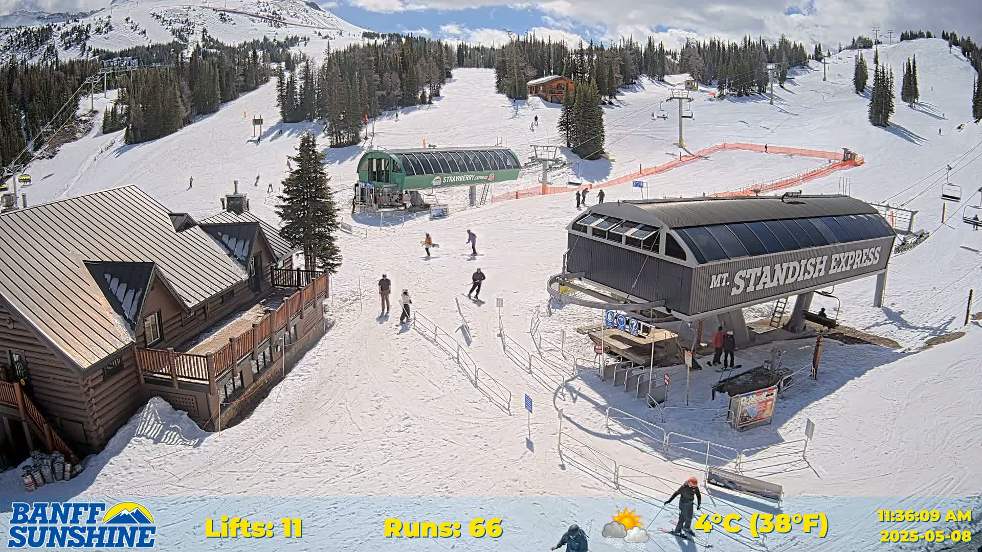 A sunny ski resort with several skiers and snowboarders on the slopes, two chairlifts, and a few log buildings.