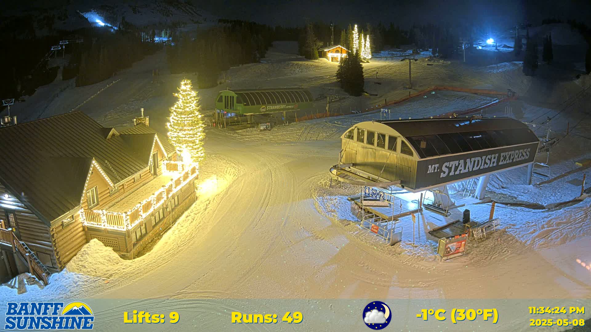 A ski resort at night, illuminated by lights on buildings and a Christmas tree, with snow on the ground and a chairlift visible under a partly cloudy sky.