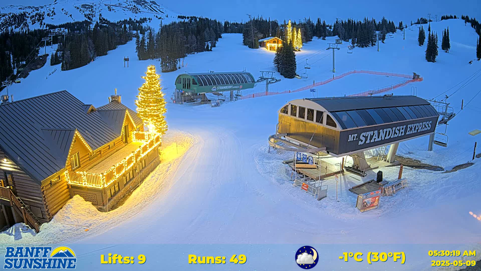 A snow-covered ski resort at night, illuminated by lights on buildings and a Christmas tree, with chairlifts and a mountain in the background, under a clear, cold sky.