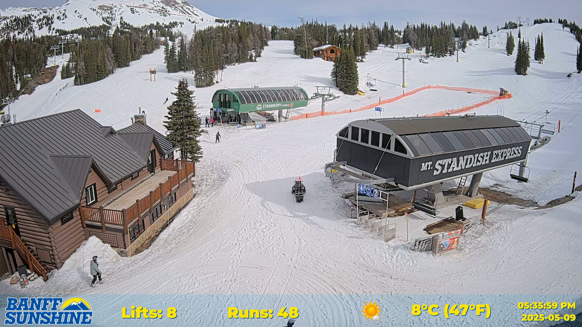 A sunny ski resort shows two chairlifts, a snow-covered lodge, and several ski runs with skiers visible on a clear, 8°C (47°F) day.
