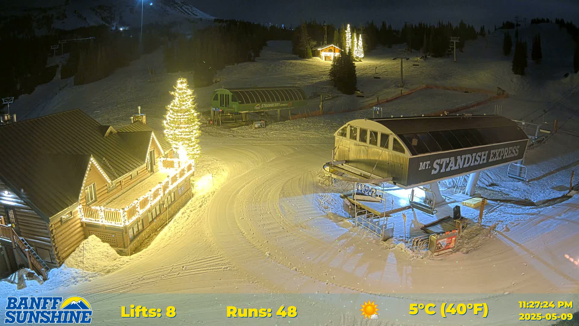 A nighttime view of a snow-covered ski resort with illuminated buildings and a ski lift, under a clear sky with a temperature of 5°C (40°F).