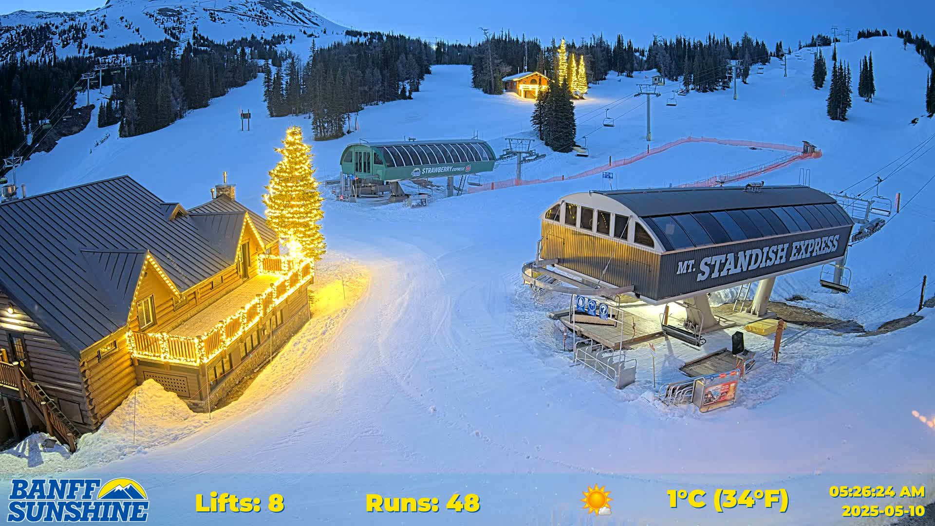 A snow-covered ski resort at twilight shows two chairlifts, a lodge with string lights, and a Christmas tree illuminated at night, with a mountain in the background.