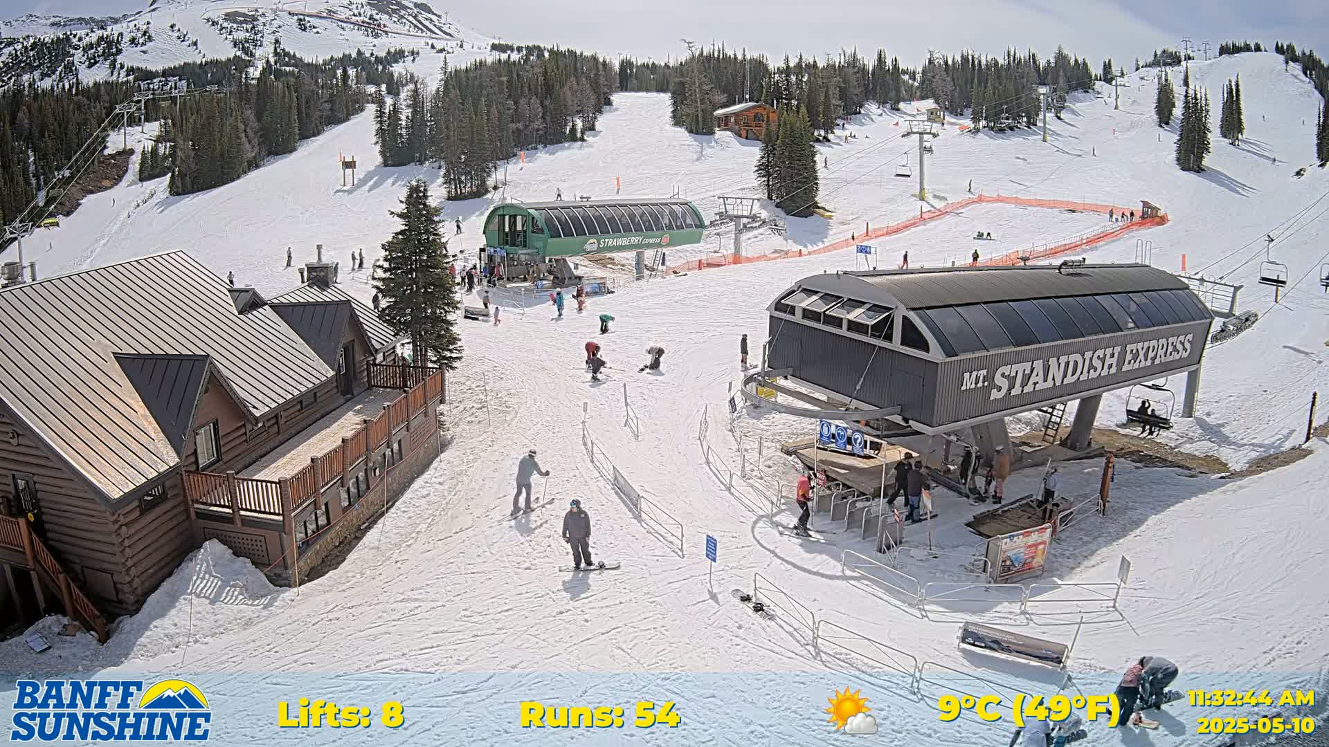 A sunny ski resort with snow-covered slopes, several chairlifts, a log cabin-style building, and skiers and snowboarders scattered on the runs.