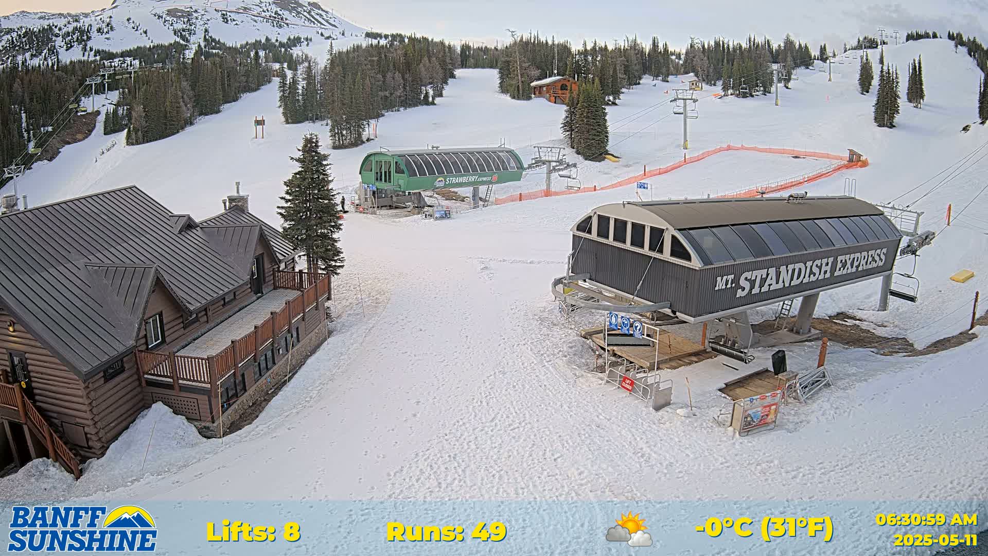 A snow-covered ski resort with two chairlifts, a log cabin, and a partly cloudy sky.