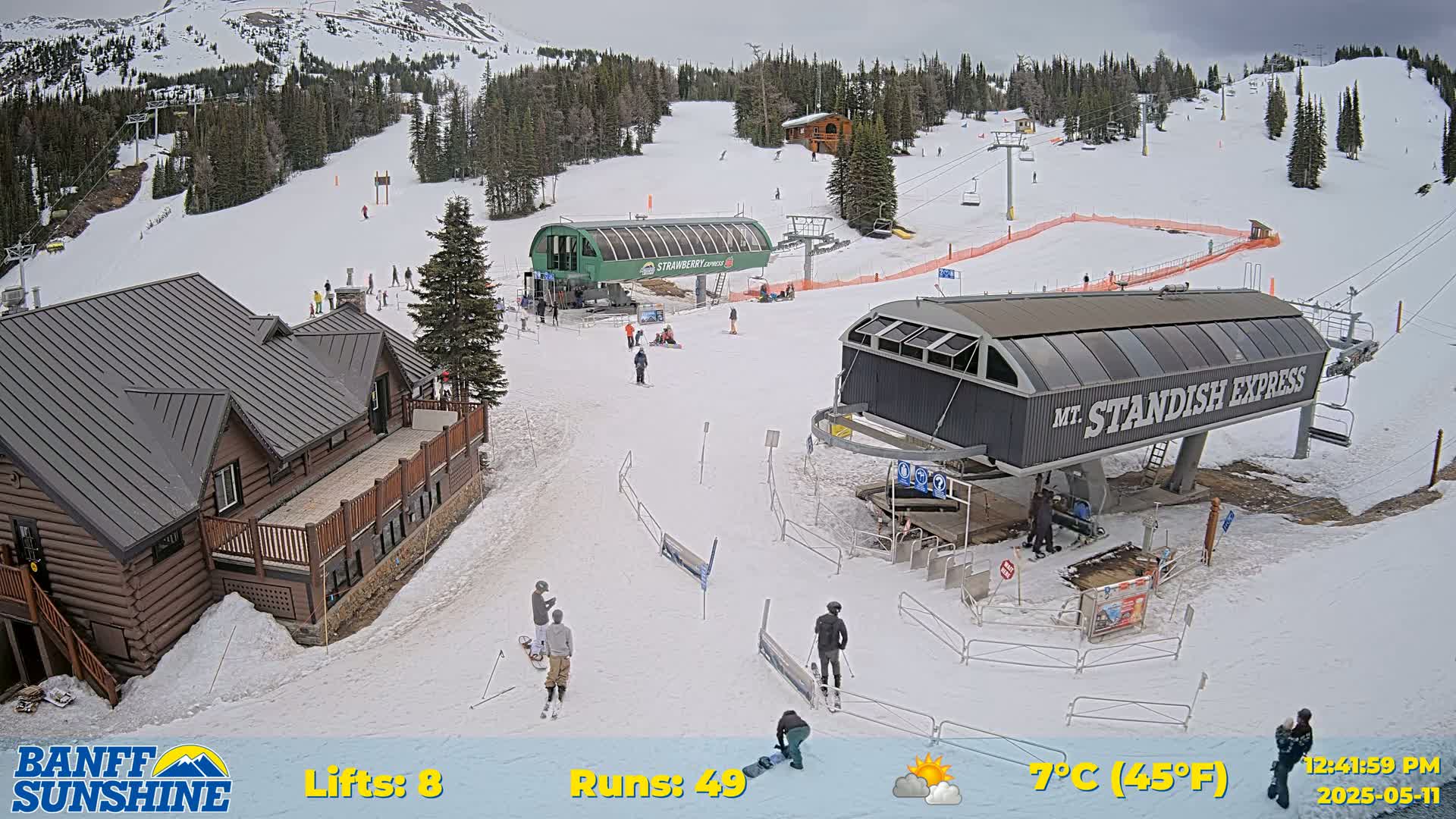 A snow-covered ski resort with two chairlifts, a few skiers and snowboarders, and a wooden lodge under partly cloudy skies.
