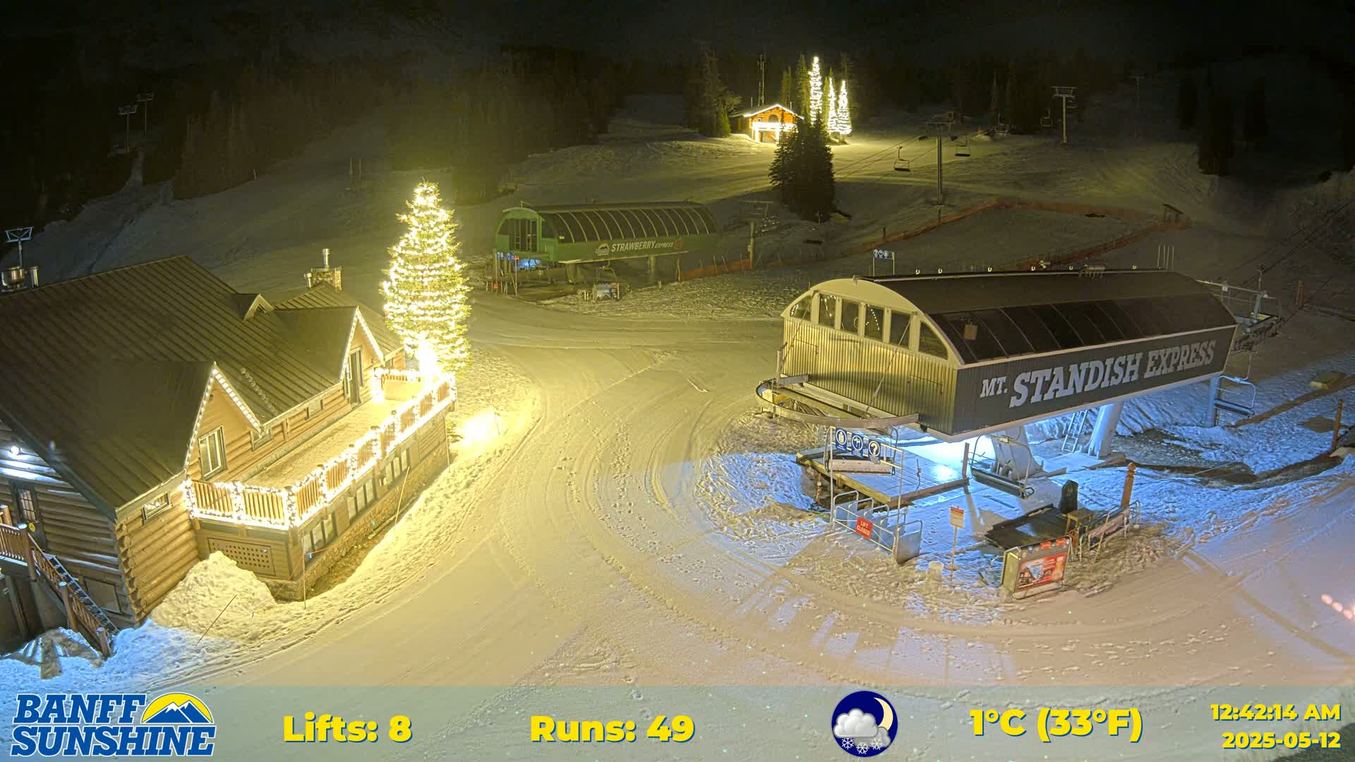 A nighttime view of a snow-covered ski resort with illuminated buildings and a chairlift, under a partly cloudy sky.