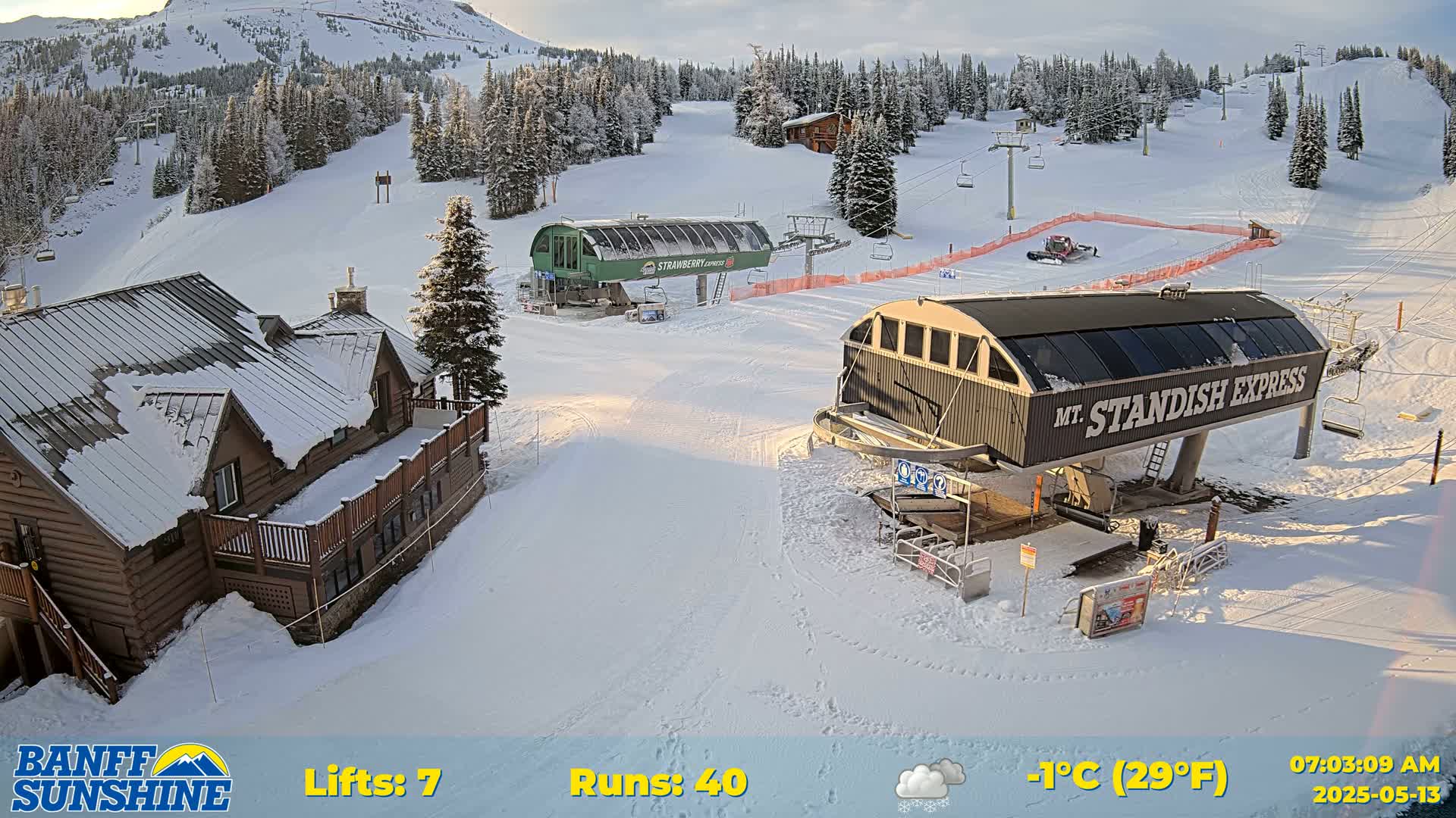 A snow-covered ski resort with two chairlifts, a lodge, and a mountain in the background under partly cloudy skies.