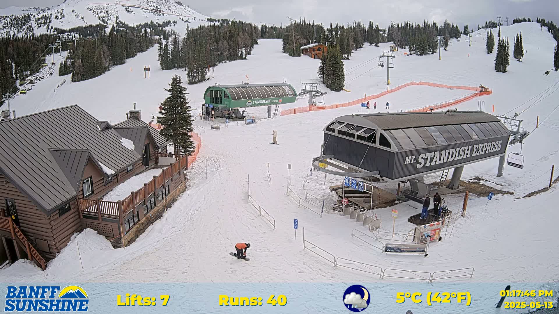 A snow-covered ski resort with two chairlifts, a lodge, and several skiers and snowboarders, under partly cloudy skies.