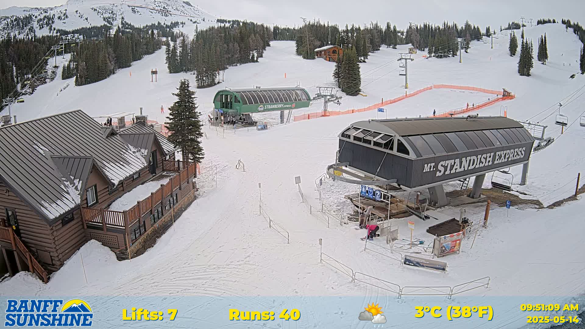 A snow-covered ski resort with two chairlifts, a wooden lodge, and several skiers visible on a partly cloudy day.