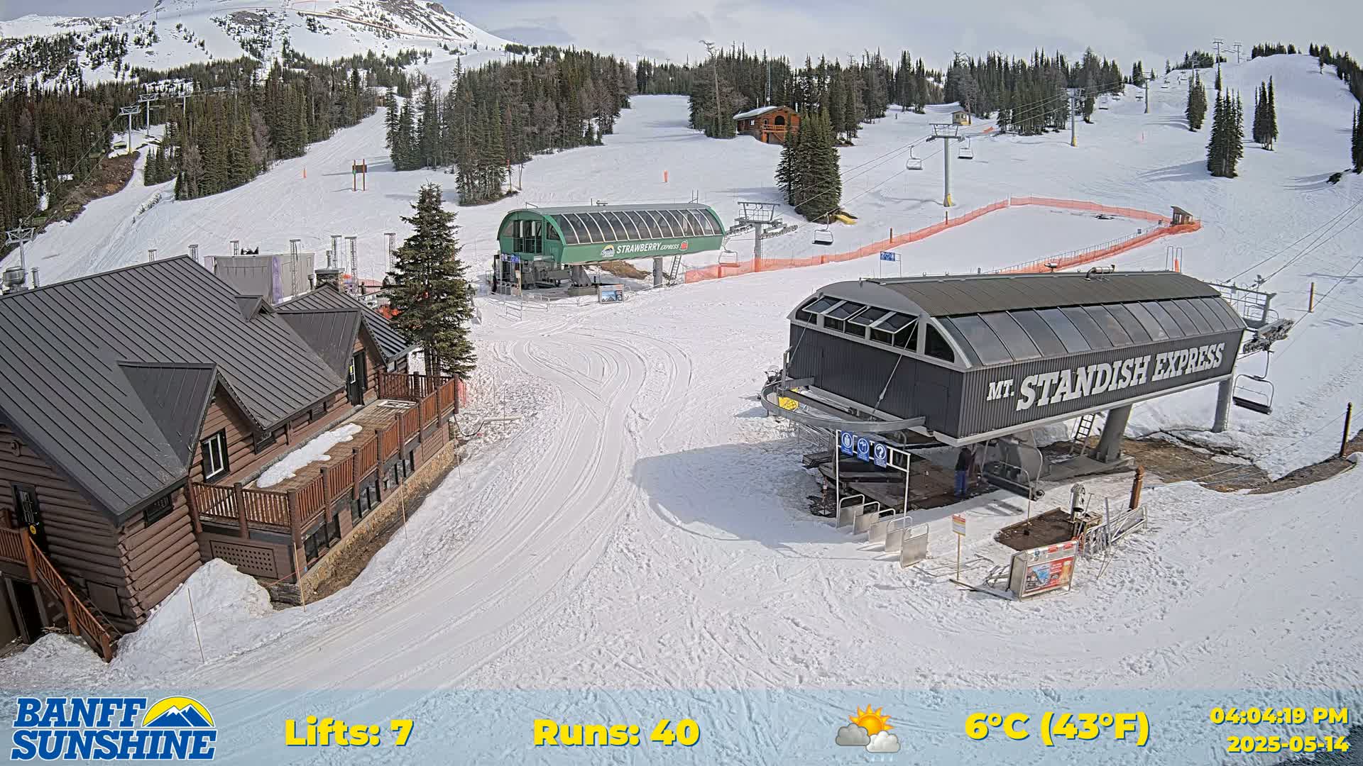 A snow-covered ski resort with two chairlifts and a log cabin, under partly sunny skies.