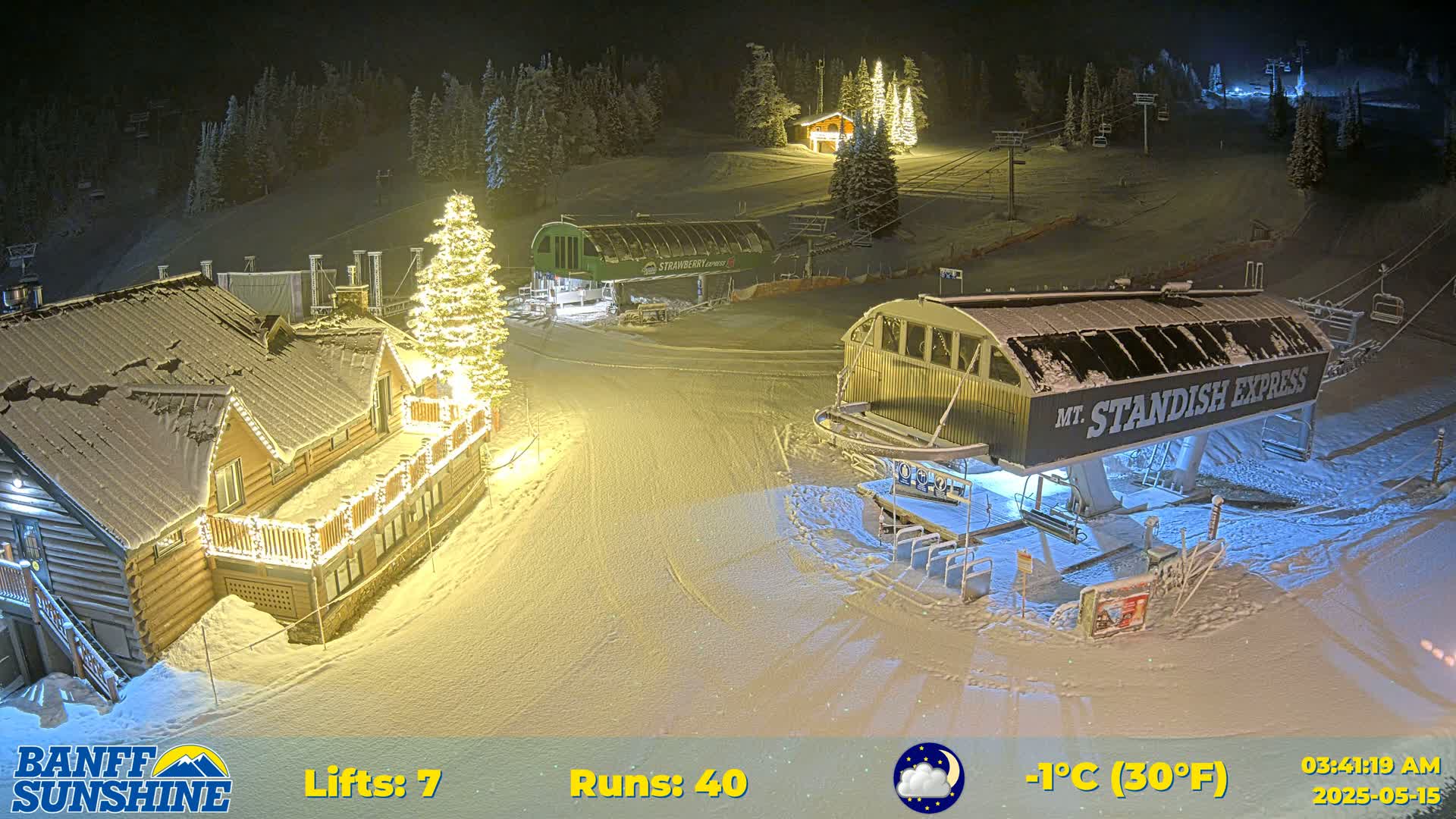 A nighttime, snow-covered ski resort scene illuminated by artificial lights, showing ski lifts, buildings, and a Christmas tree.