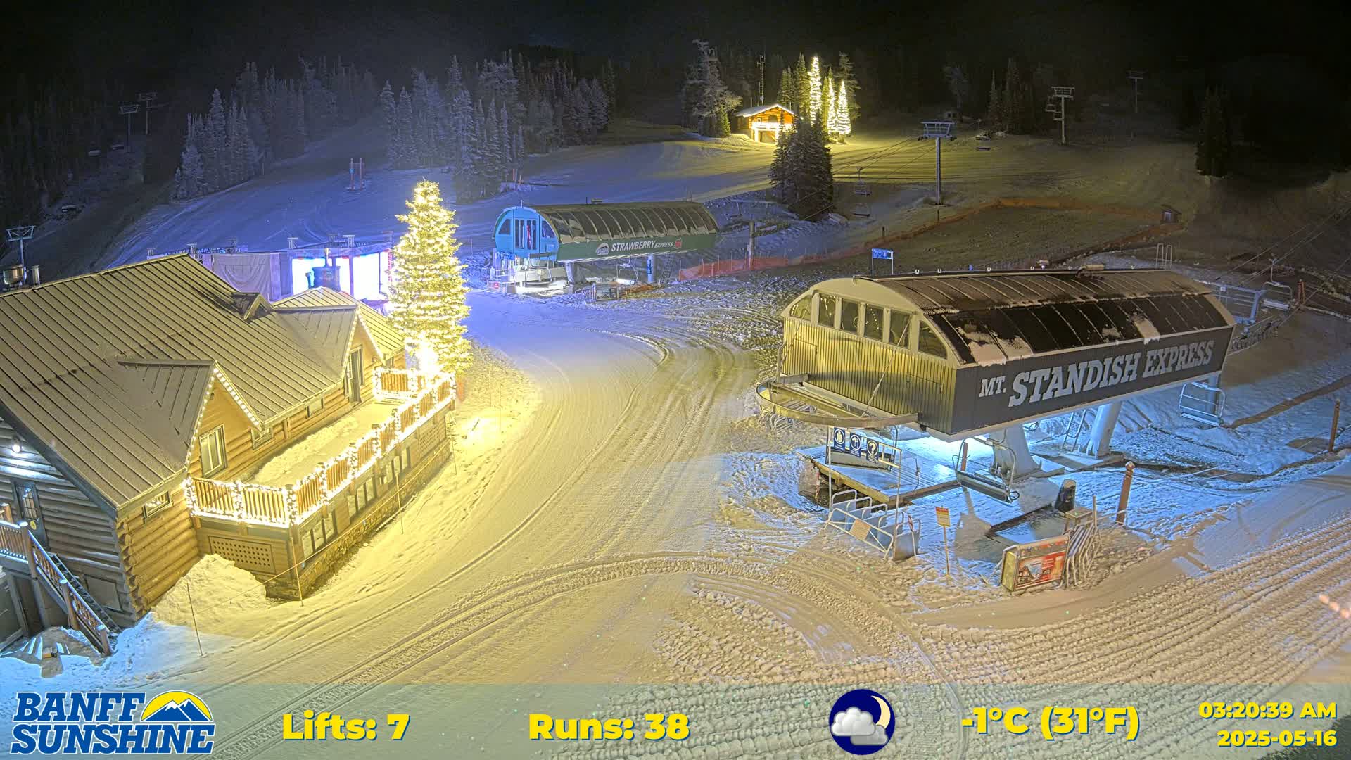 A snow-covered ski resort at night, illuminated by lights on buildings and a Christmas tree, with a chairlift visible under partly cloudy skies.