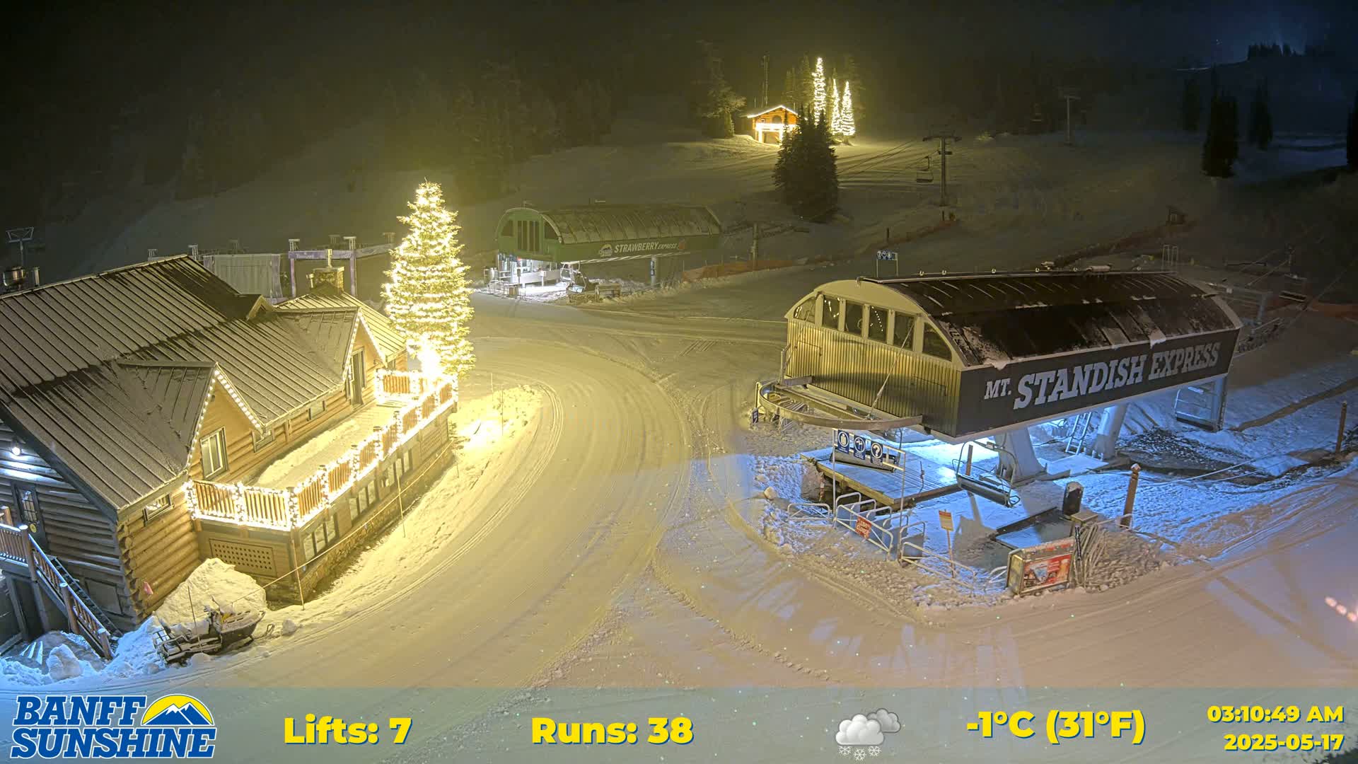 A snow-covered ski resort at night, illuminated by lights on buildings and a Christmas tree, with several ski lifts visible under a partly cloudy sky.