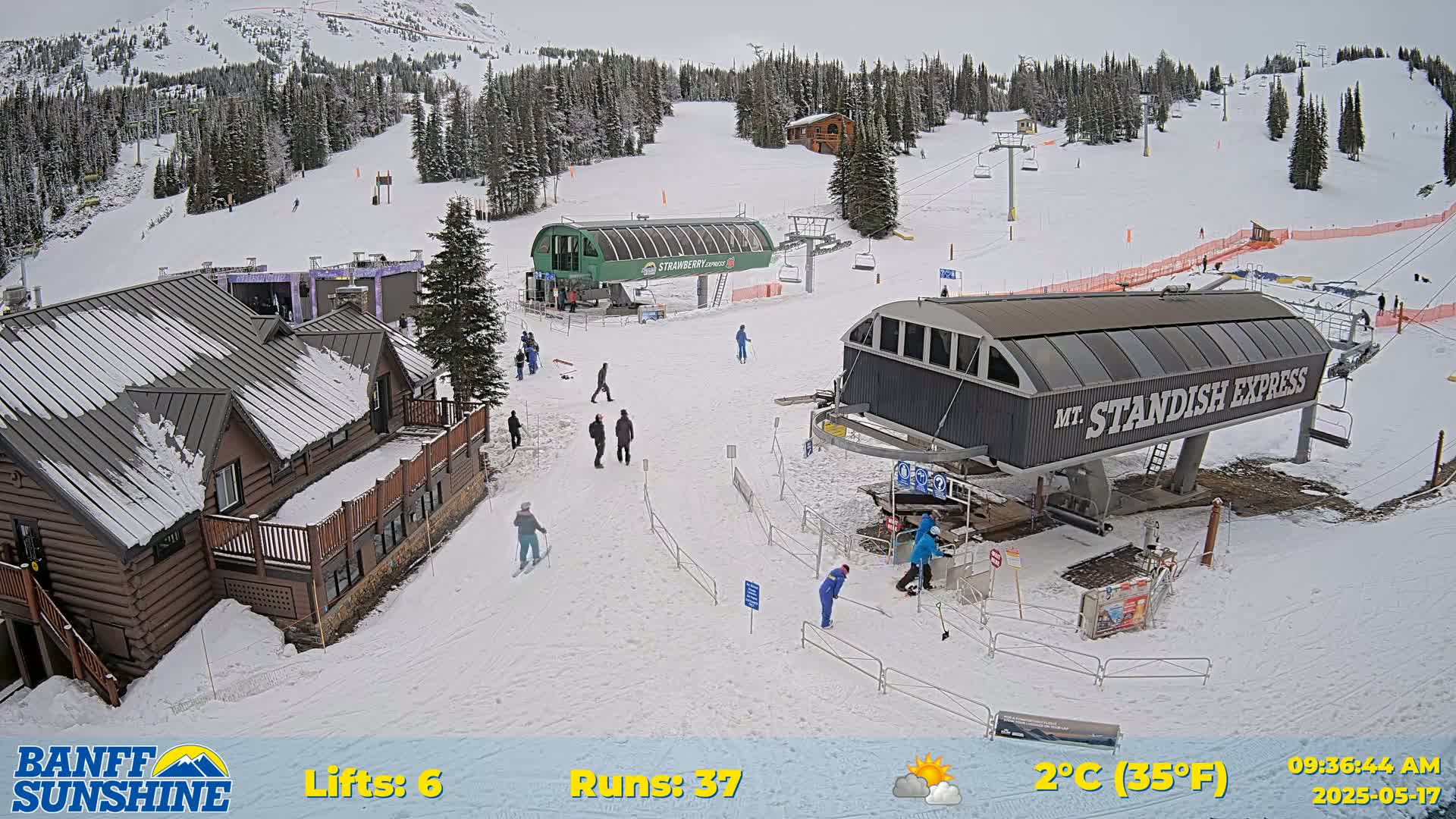 A snowy ski resort scene shows several skiers and snowboarders near two chairlifts and a lodge under partly cloudy skies.