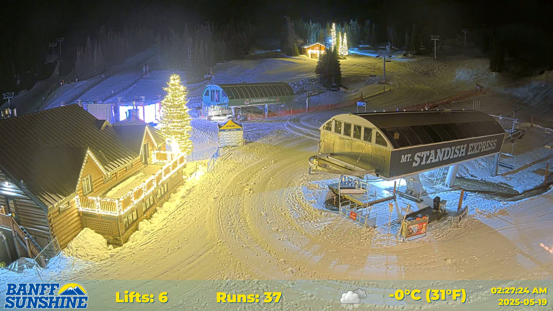 A nighttime view of a snow-covered ski resort with illuminated buildings and ski lifts, under a partly cloudy sky at 0°C (31°F).