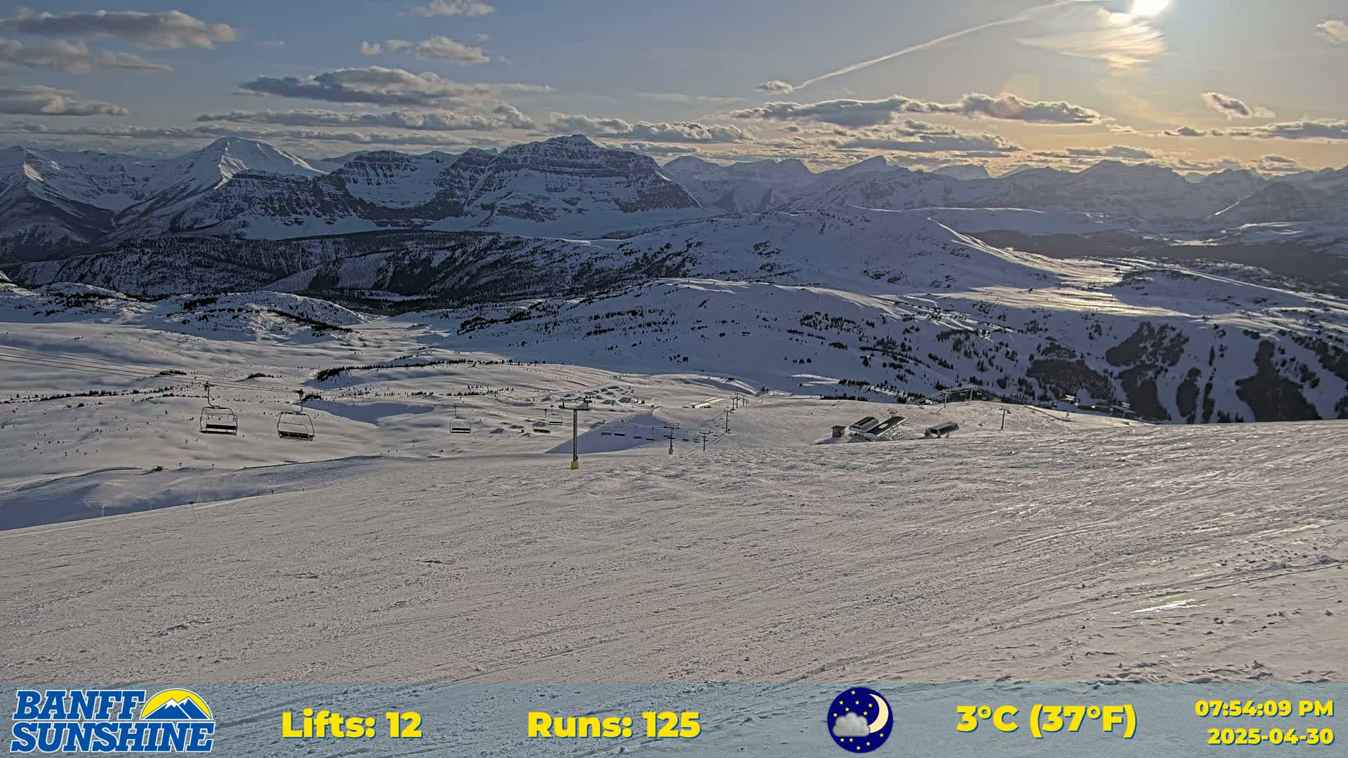 A snow-covered ski resort with chairlifts is nestled in a valley, overlooking a vast mountain range under a partly cloudy sunset.