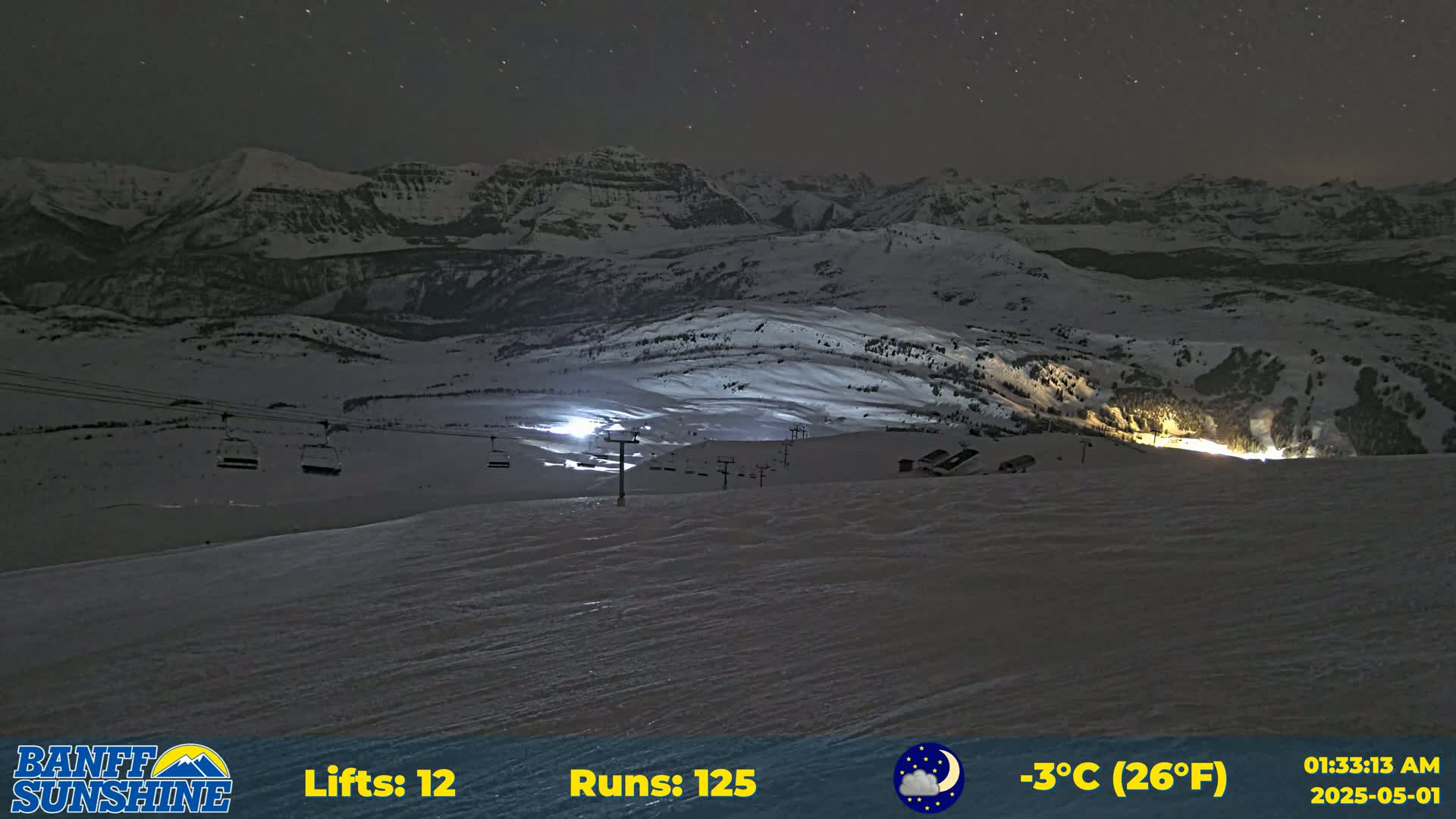 A nighttime view of a snow-covered ski resort illuminated by lights, with a mountain range in the background under a partly cloudy sky.