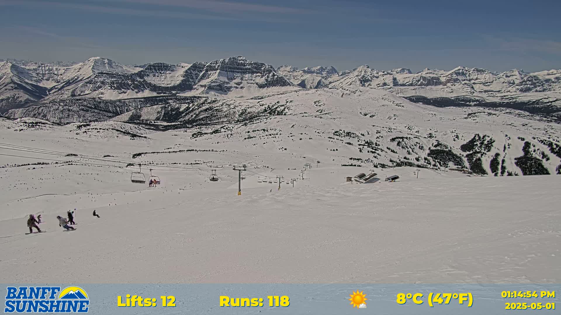 A sunny ski slope with several skiers and chairlifts is nestled among snow-covered mountains.