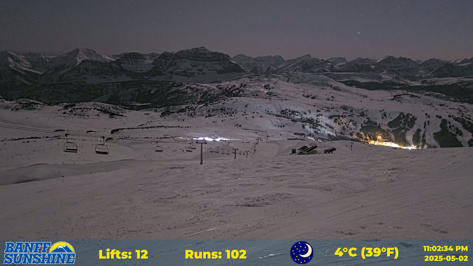 A snow-covered ski resort at night under a dark sky, with several chairlifts visible and mountains in the background.