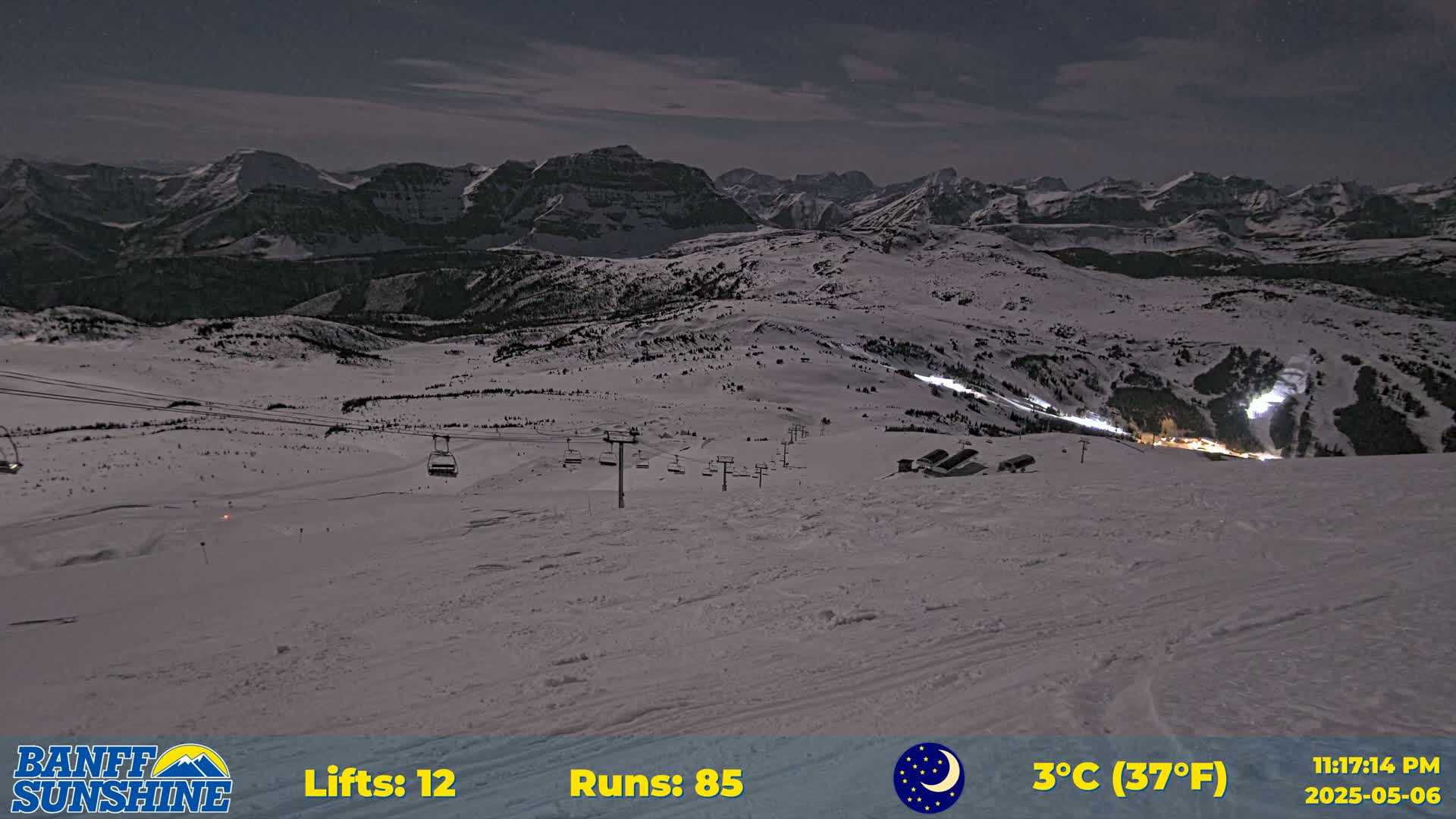 A snow-covered ski resort at night under a mostly clear sky, with several chairlifts visible and mountains in the background.