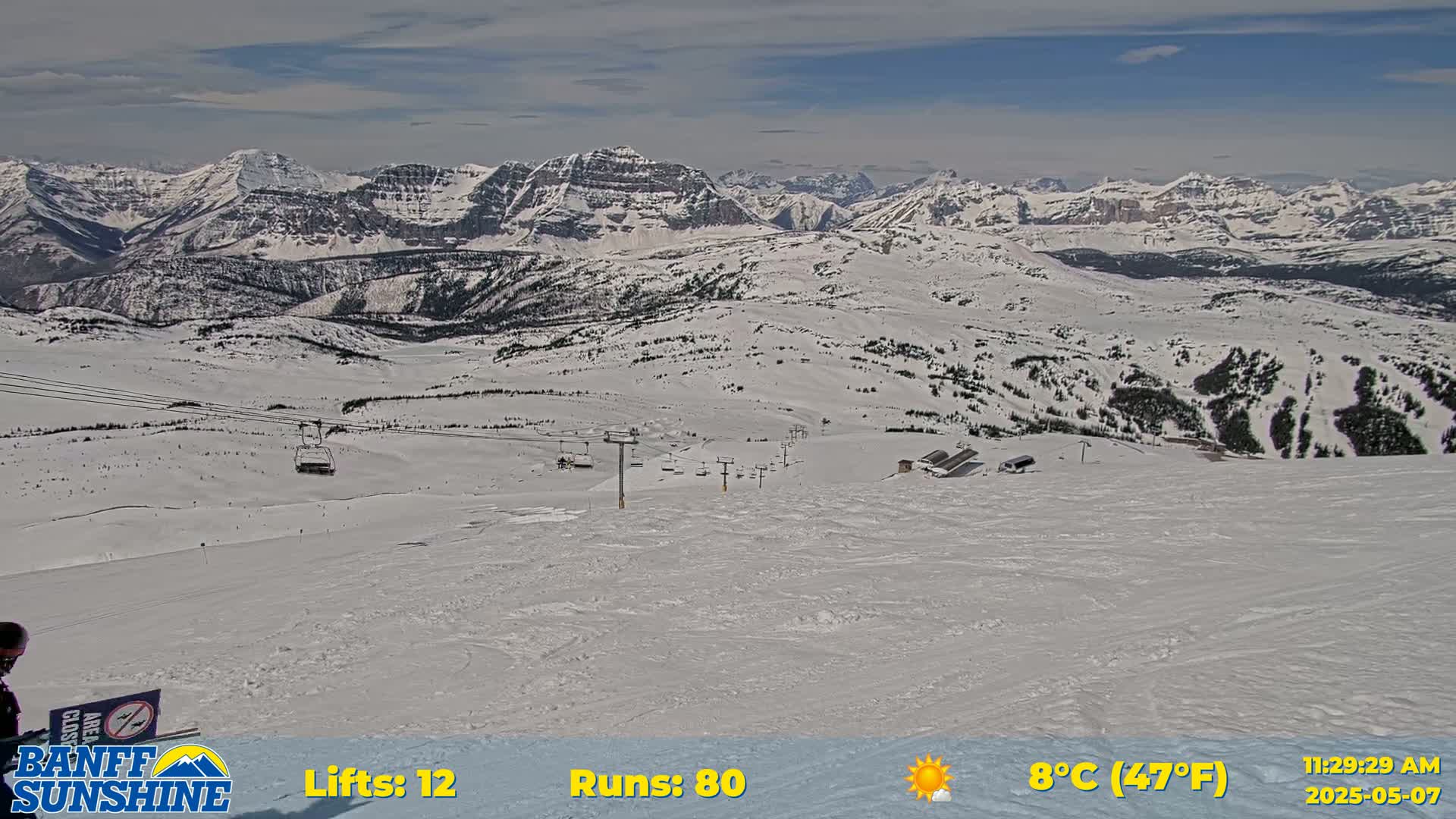 A snow-covered ski resort with chairlifts and mountain ranges in the background on a partly sunny day.