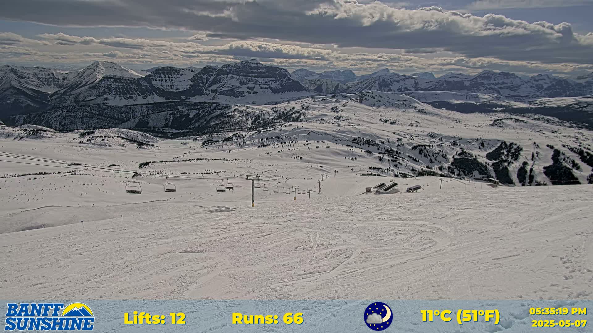 A mostly cloudy day reveals a snowy mountain ski resort with chairlifts and several ski runs, overlooking a vast mountain range.