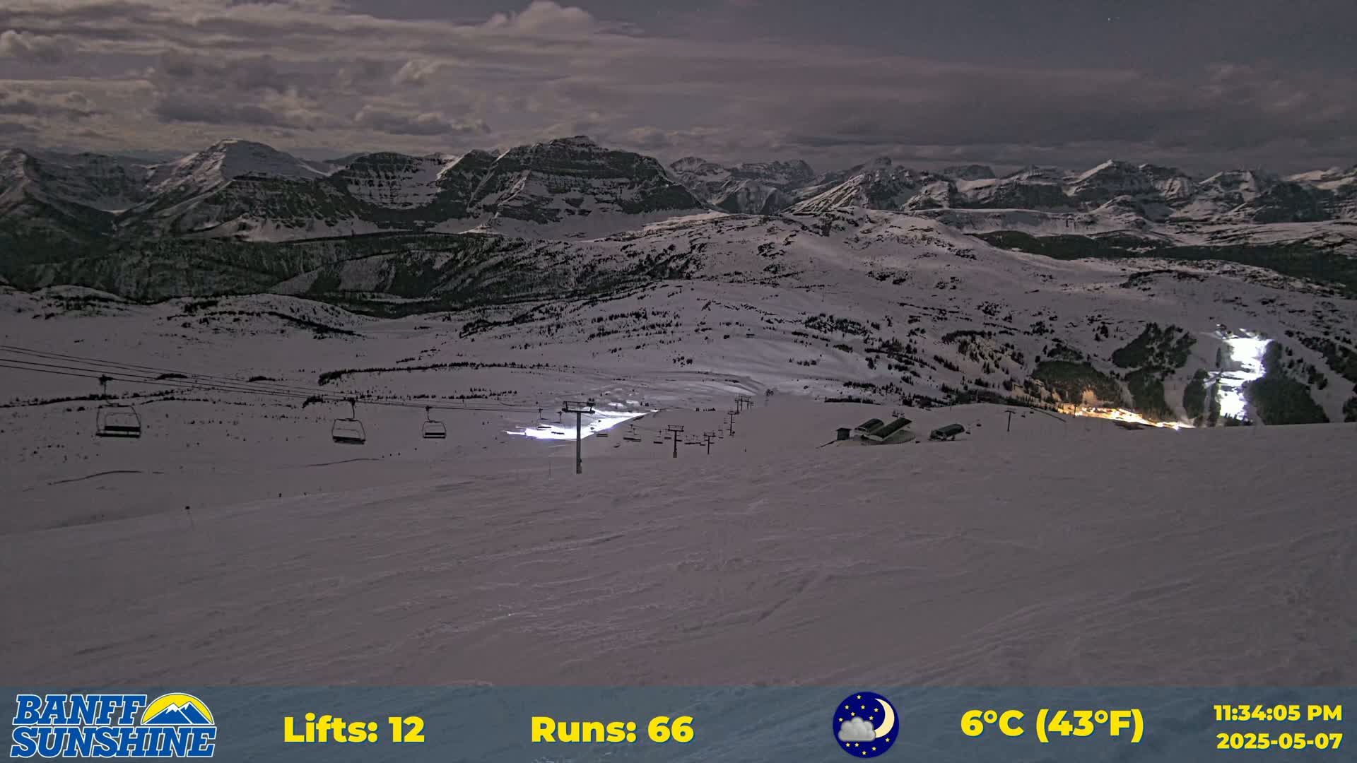 A nighttime view of a snow-covered ski resort with chairlifts and illuminated slopes under a partly cloudy sky.