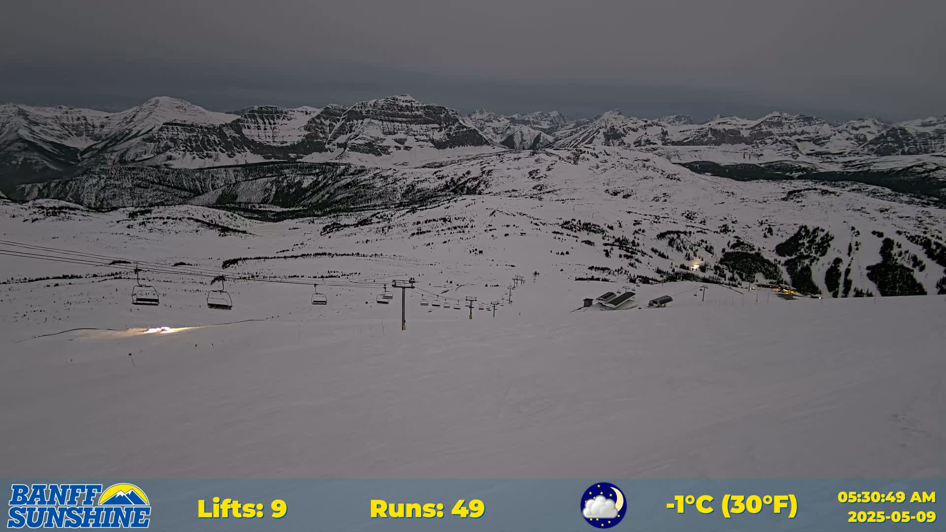 A snow-covered ski resort at night under a dark, overcast sky, showing several chairlifts and ski runs against a backdrop of snow-capped mountains.