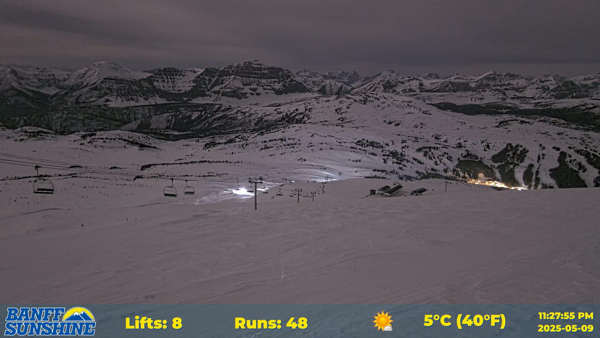 A snow-covered ski resort at night, under a partly cloudy sky, shows several chairlifts and illuminated ski runs against a backdrop of snow-capped mountains.