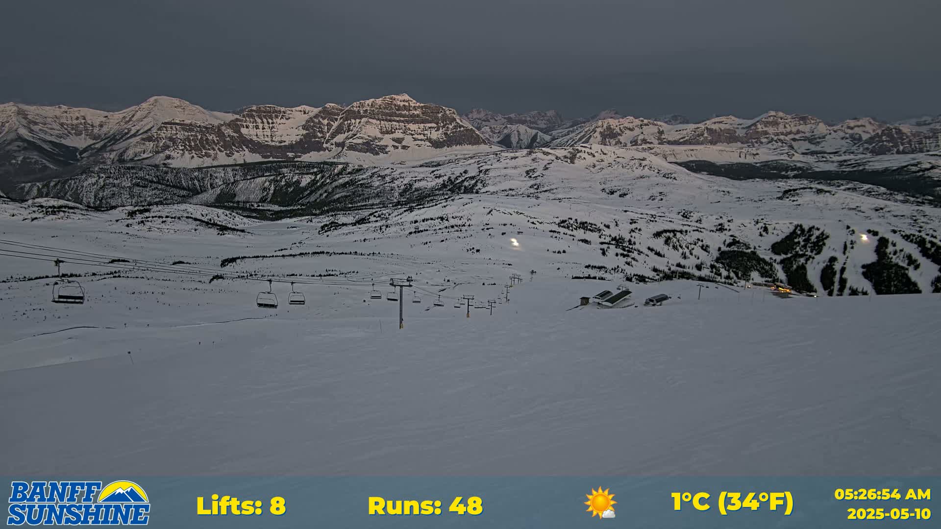 A snowy mountain landscape at dusk shows several ski lifts and runs, with a mountain range in the background under a cloudy sky.