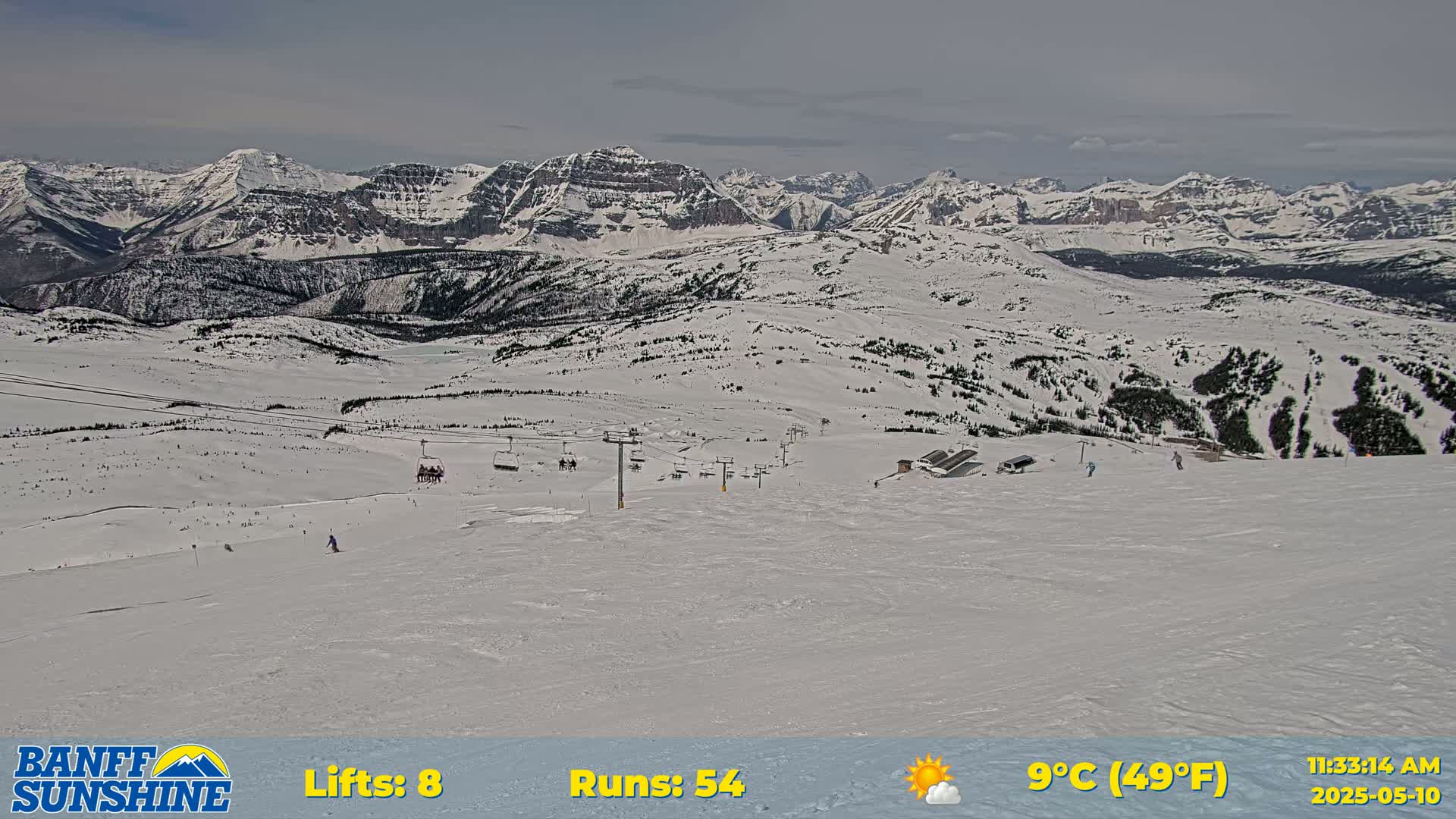 A snowy mountain ski resort with mostly sunny skies shows several ski runs and chairlifts against a backdrop of snow-covered mountains.