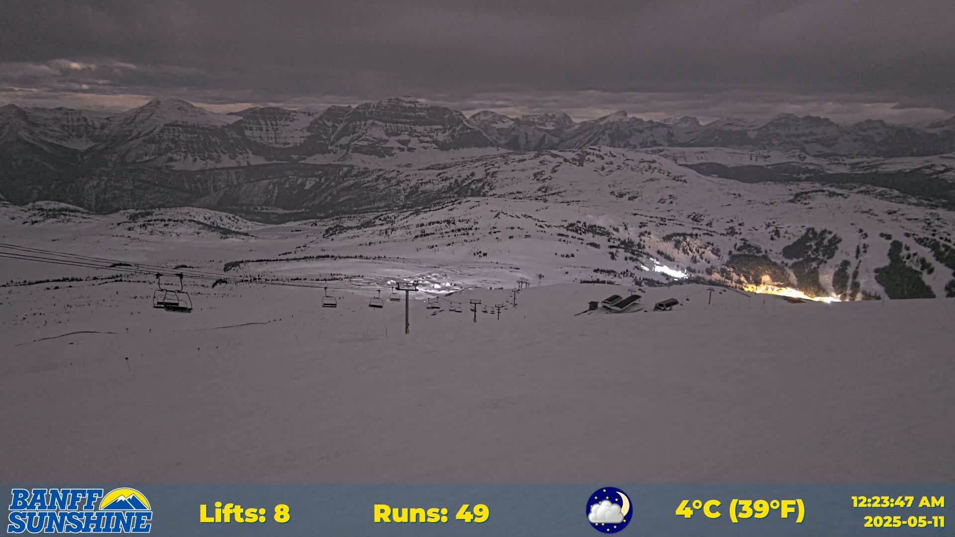 A snow-covered ski resort at night under a cloudy sky, with several chairlifts visible and lights illuminating the base area.