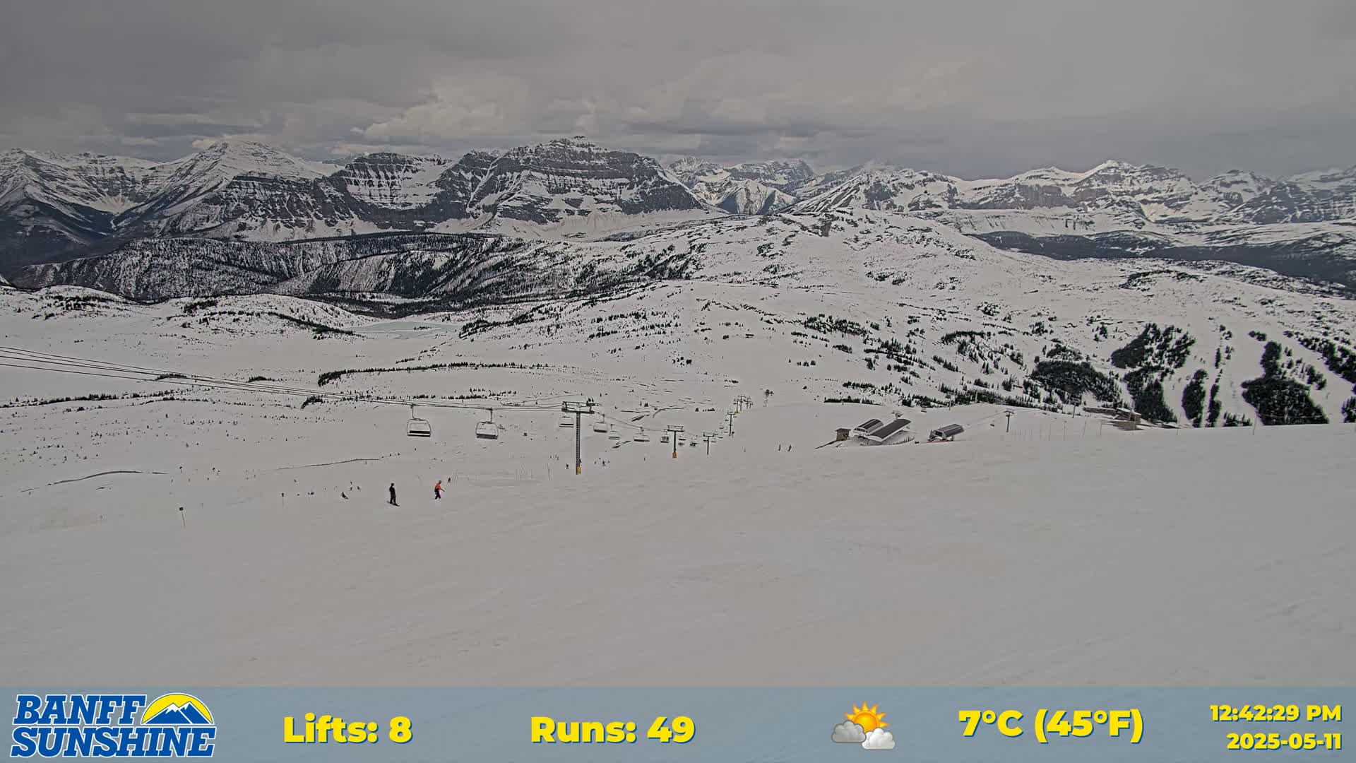 A snowy mountain ski resort scene under an overcast sky shows several chairlifts and skiers on a slope with distant snow-covered mountains in the background.