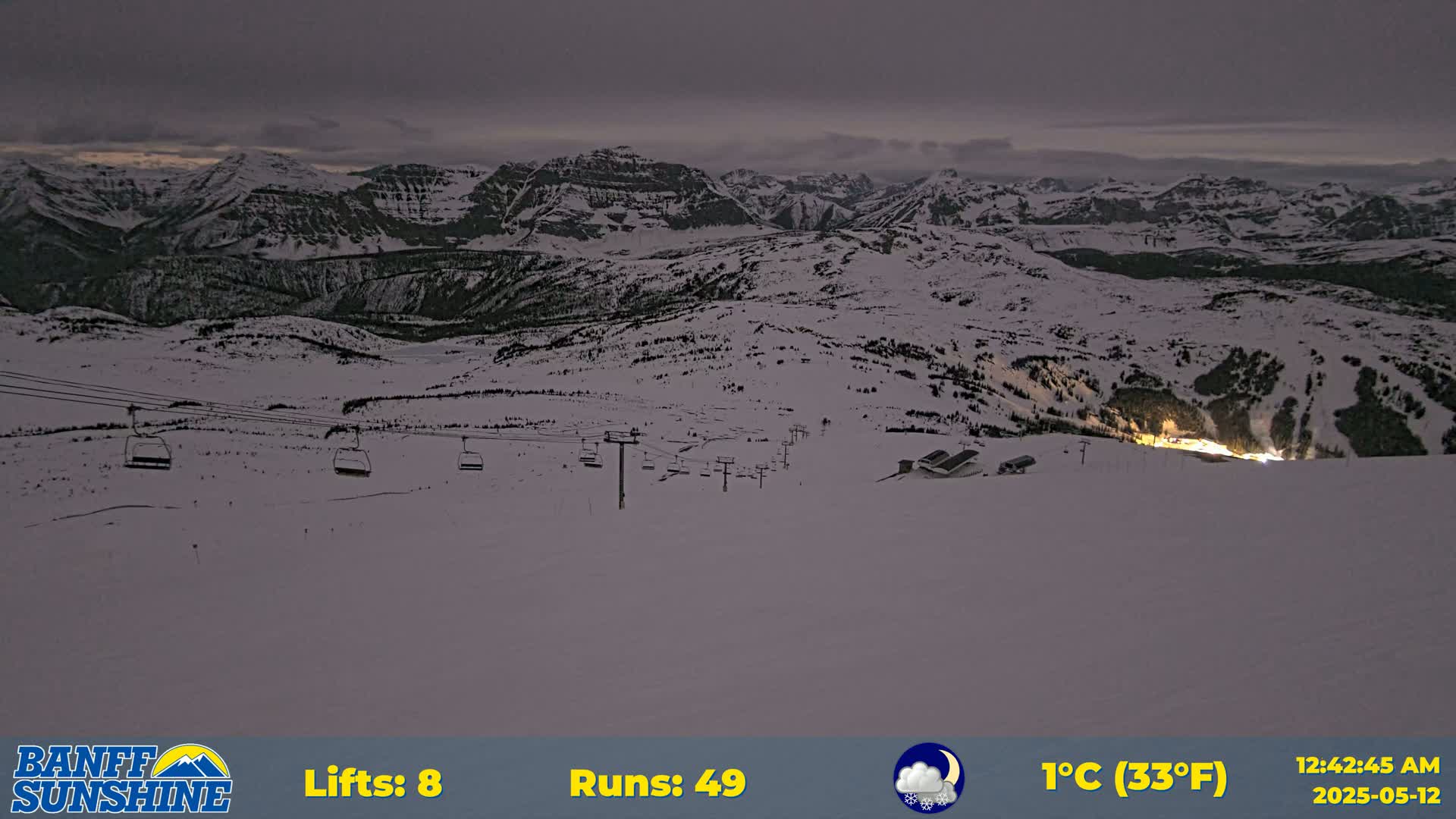 A snow-covered mountain ski resort at night under a cloudy sky, showing several ski lifts and illuminated buildings in the distance.