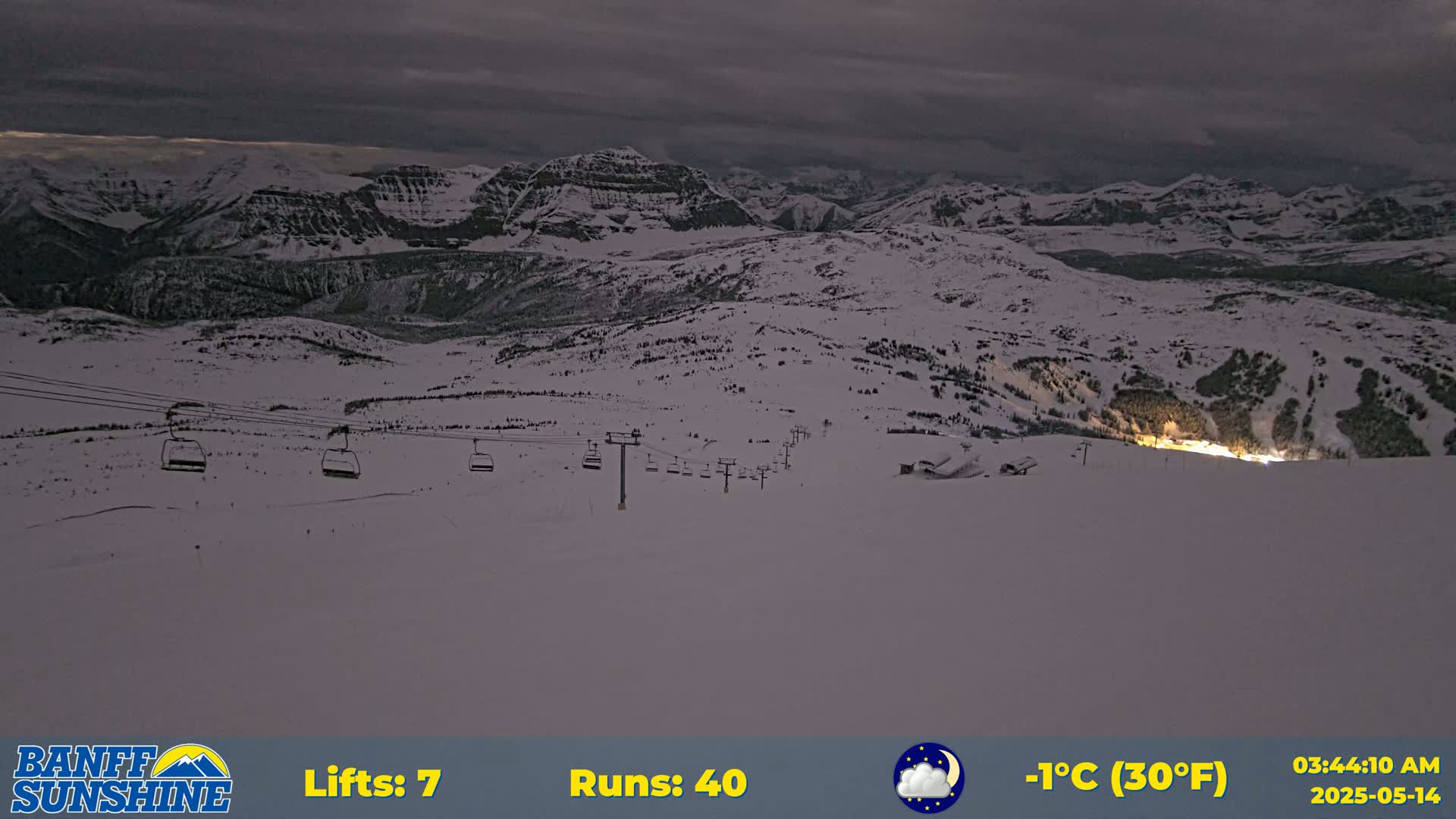 A nighttime view of a snow-covered ski resort with several chairlifts and mountain ranges in the background under a cloudy sky, with a temperature of -1°C (30°F).