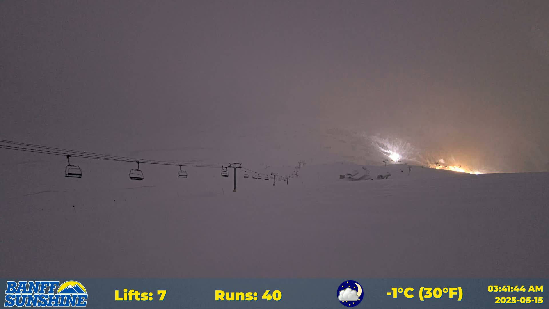 A snow-covered ski slope at night, illuminated by lights in the distance, shows a chairlift with several empty chairs, under a hazy, overcast sky.