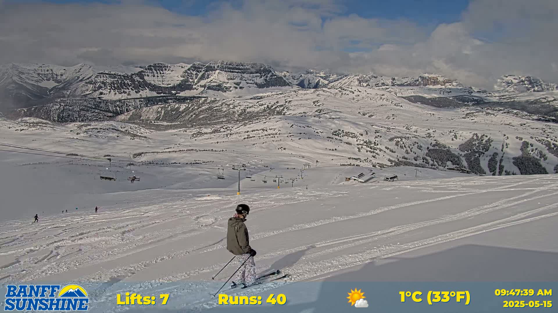 A skier stands on a snowy mountain slope overlooking a valley with snow-covered mountains under partly cloudy skies.