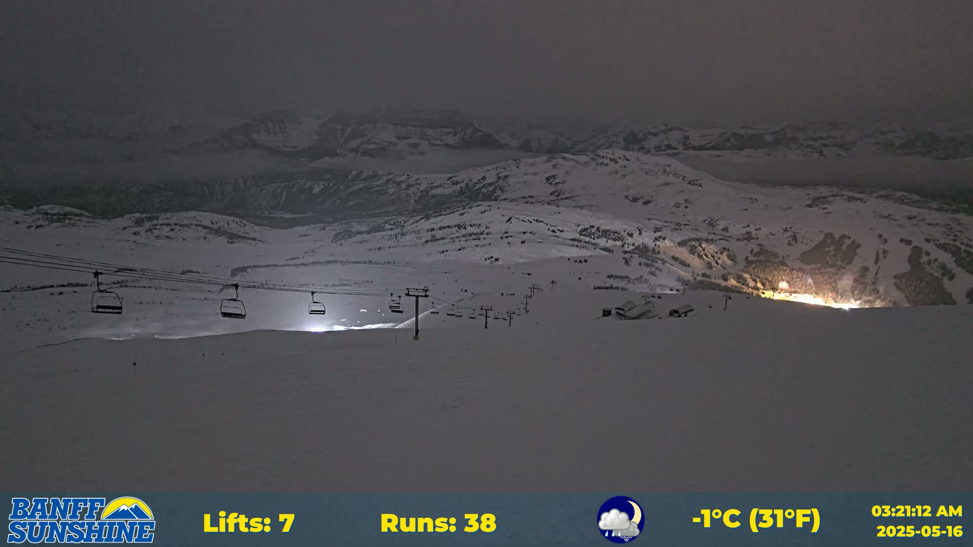 A nighttime view of a snow-covered ski resort with several chairlifts and illuminated areas, under a cloudy, cold sky.