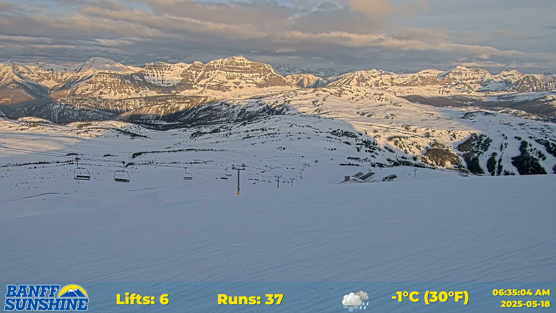 A snow-covered ski resort with several chairlifts sits below a mountain range under a partly cloudy sky, lightly snowing.